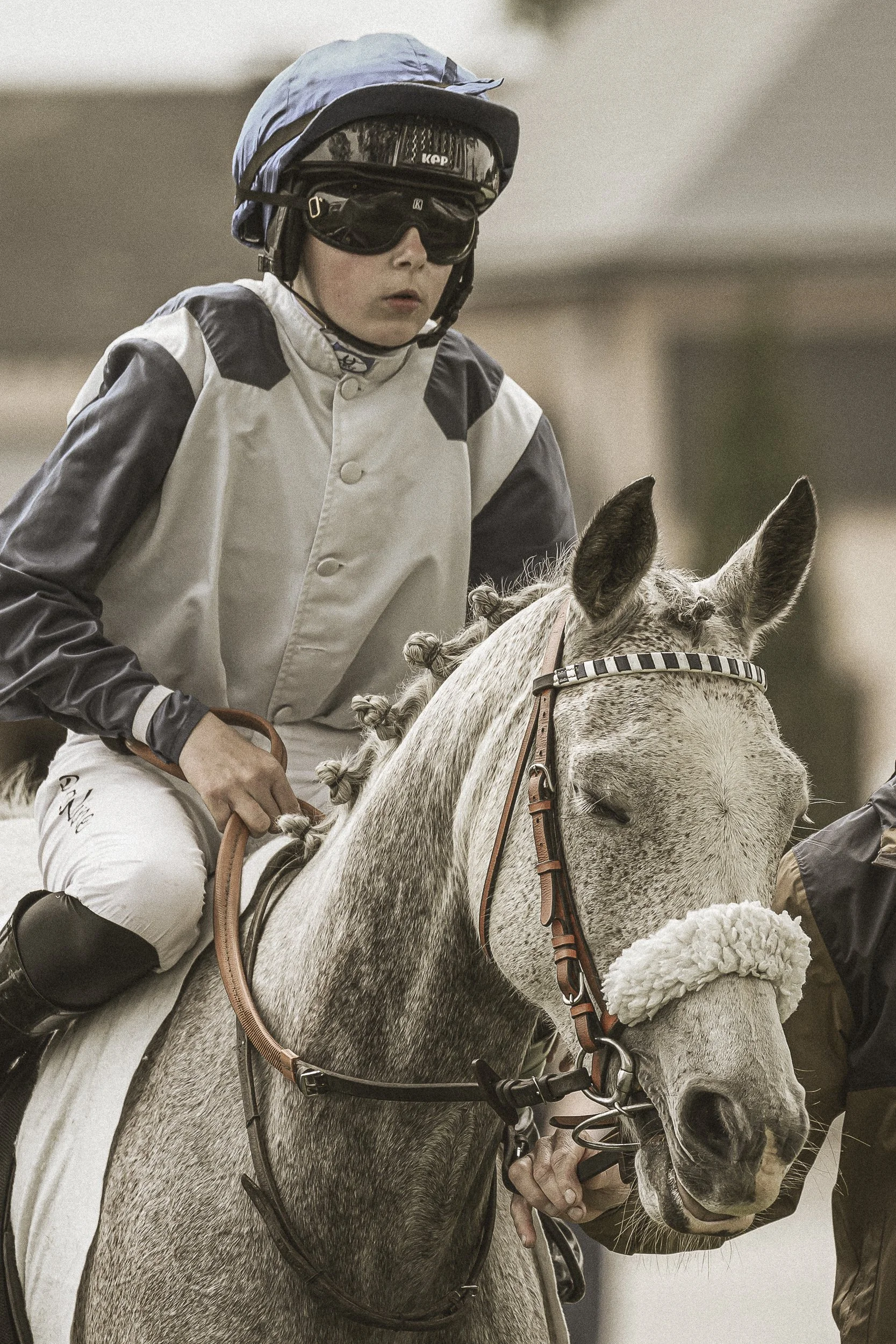 Jockey féminin portant casque et lunettes, à cheval avec une selle blanche et un masque de bride, dans une compétition équestre.
