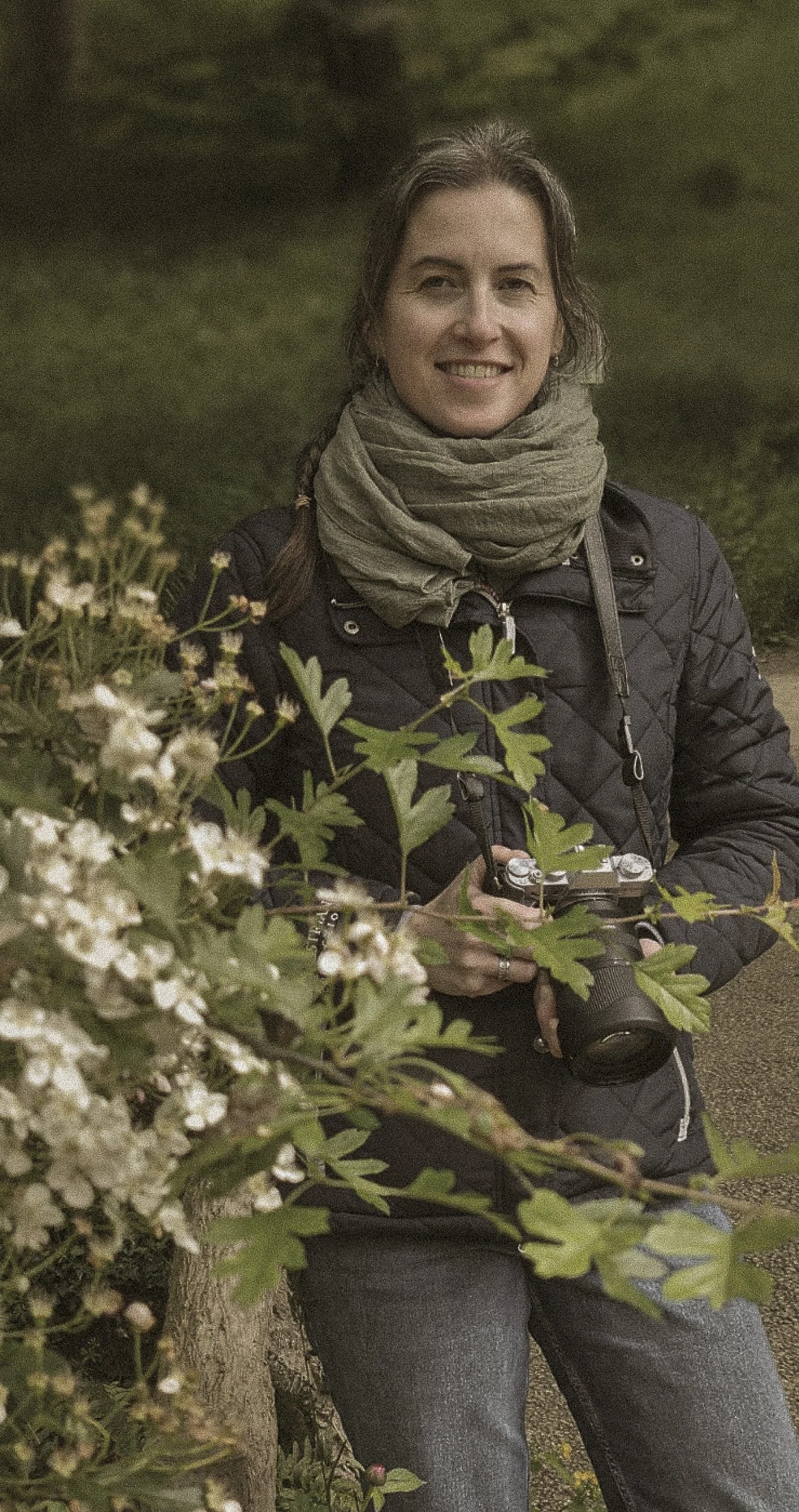 Femme tenant un appareil photo, souriant, vêtue d'une veste noire et d'une écharpe beige, entourée de végétation.