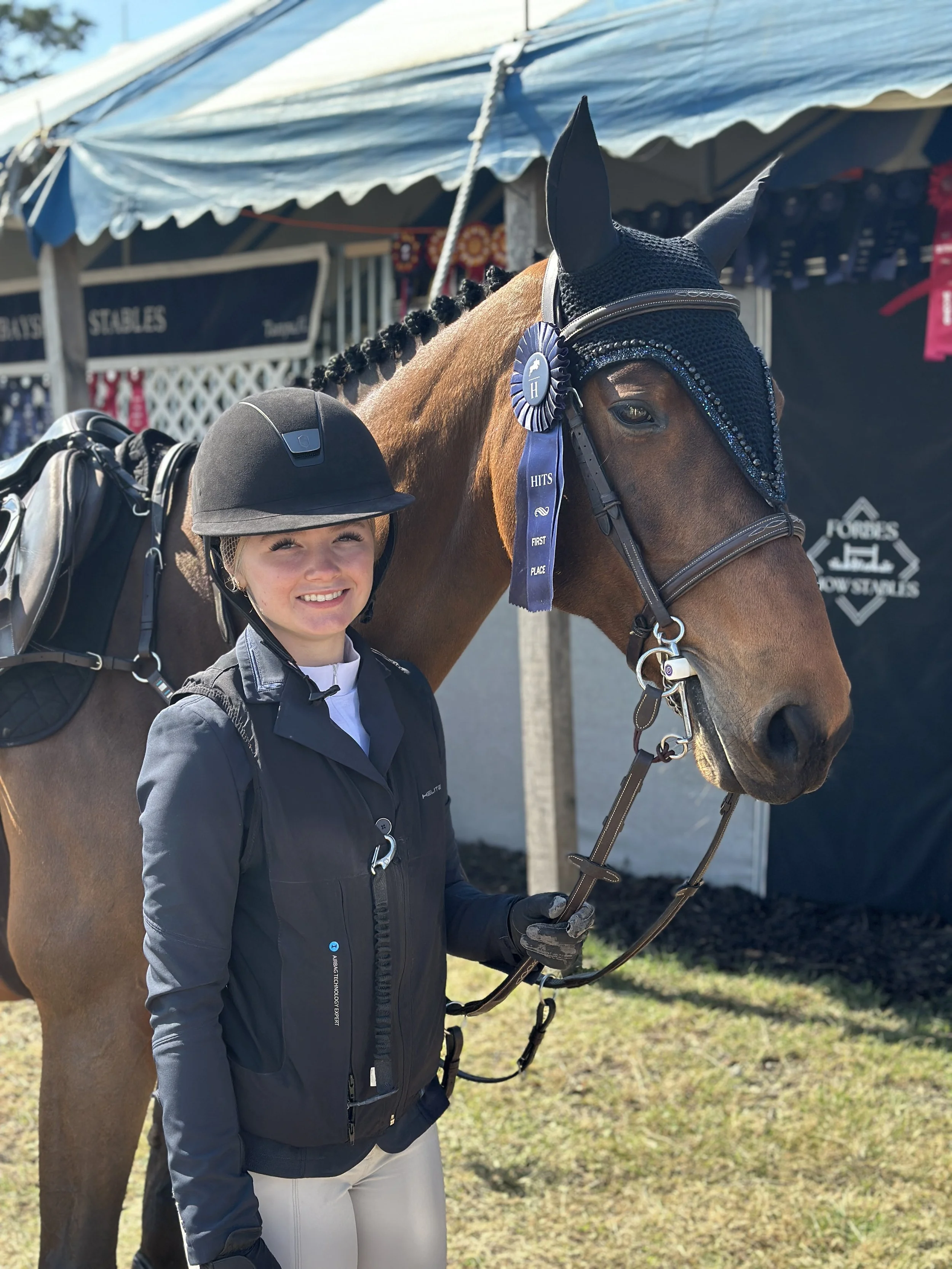 A young woman with blonde hair in braids, wearing a black equestrian helmet, standing next to a brown horse with a black headgear. The woman is smiling, dressed in a black riding jacket, white blouse, beige riding pants, and black gloves, holding the