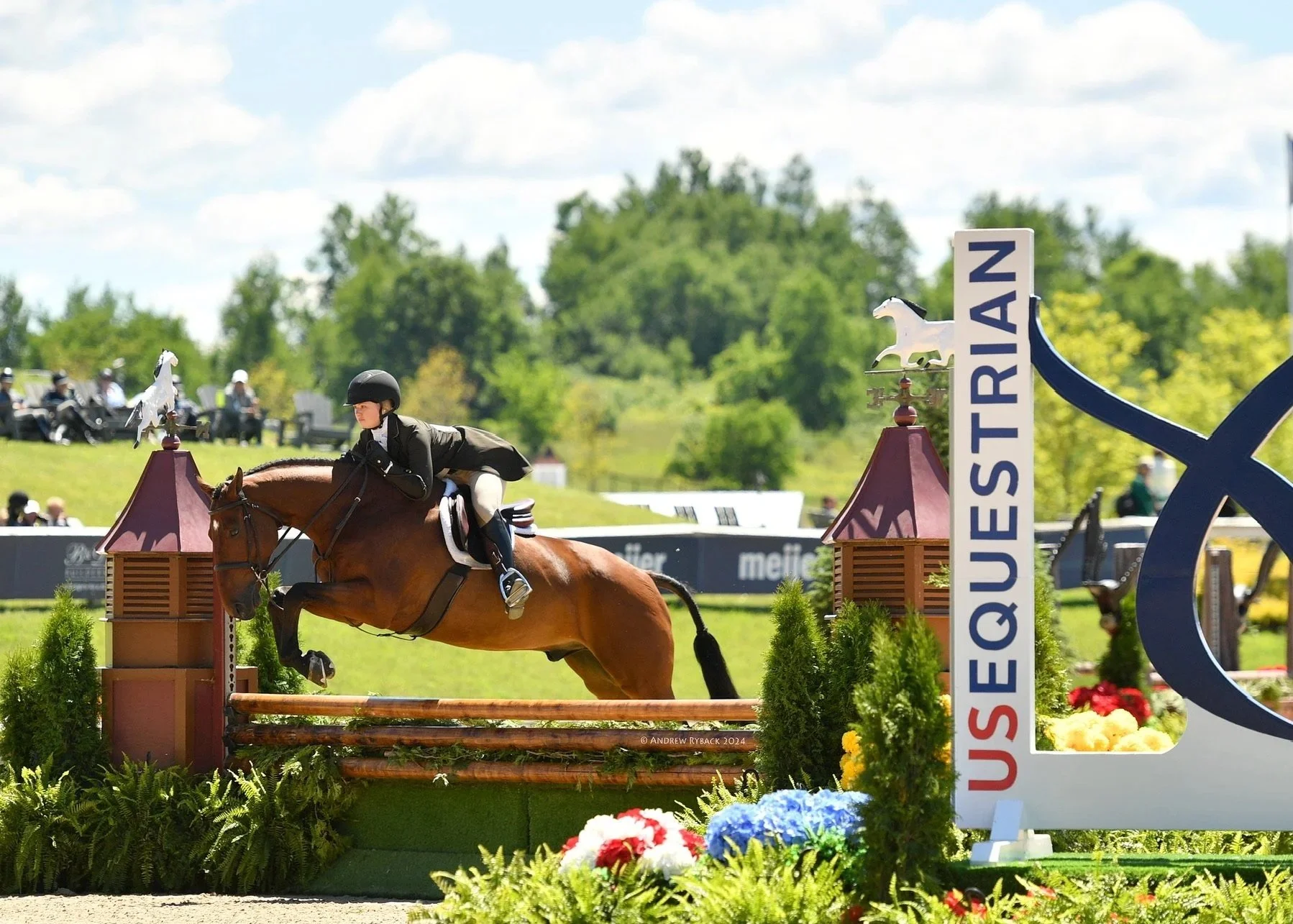 A rider in equestrian attire riding a horse over a jump at a show. The jump includes a blue sign with the word "QUESTRIAN" and decorative elements, with American and regional flags in the background under a partly cloudy sky.