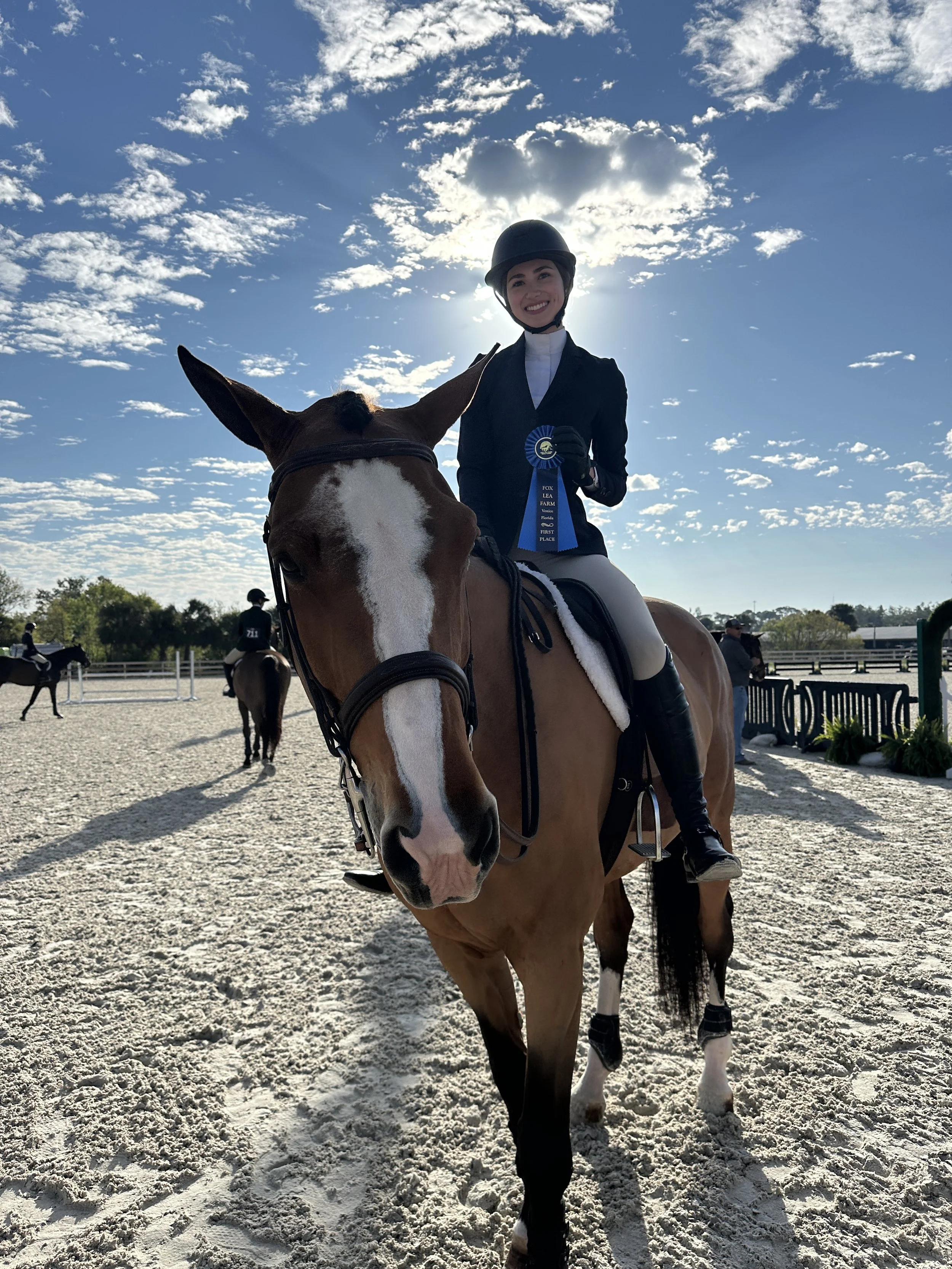 Young woman in riding gear, wearing a black helmet, smiling while riding a brown and white horse with a white stripe on its face, at an equestrian event with a blue sky and scattered clouds in the background.