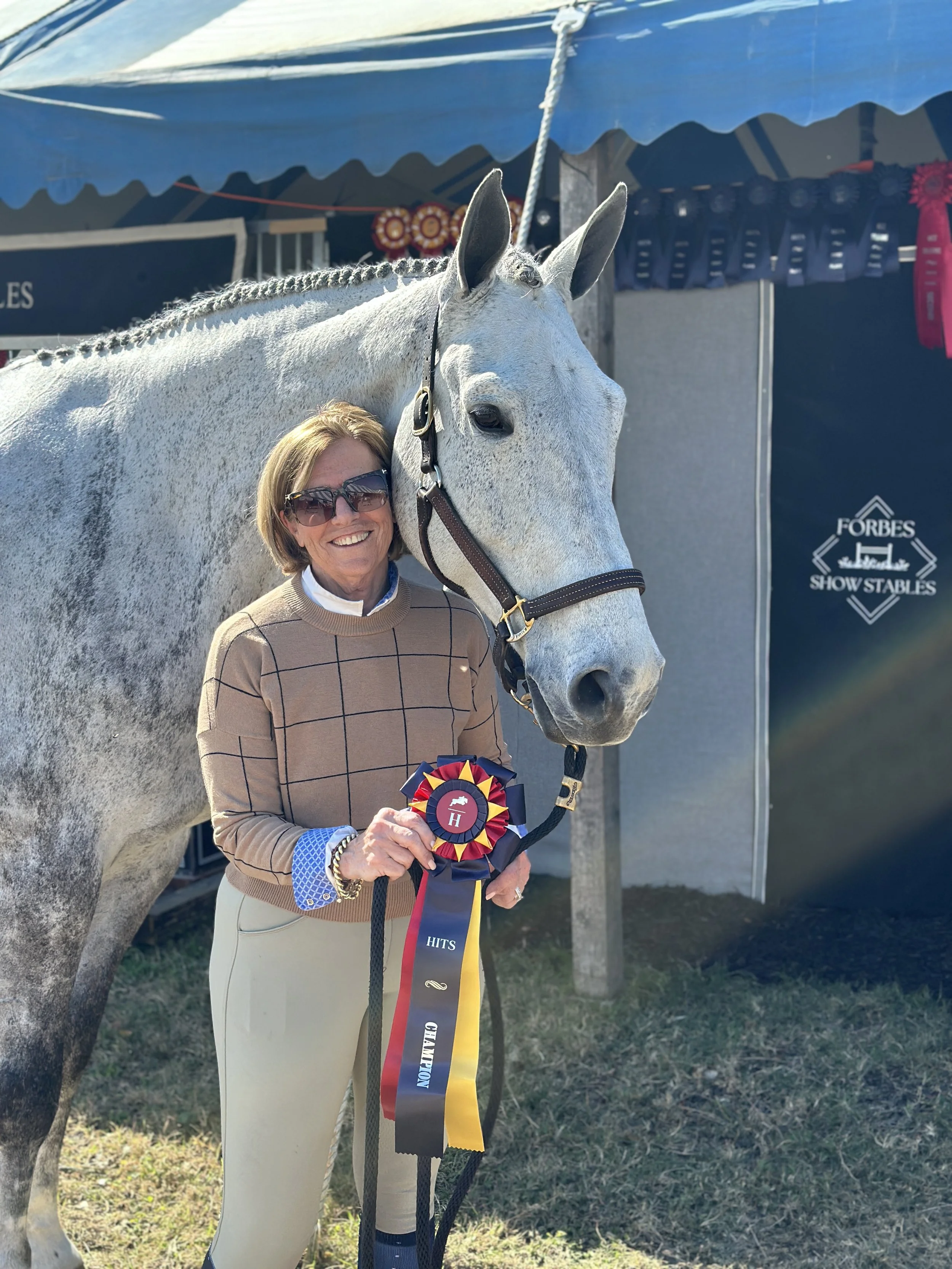 A woman with short blonde hair, wearing sunglasses, a tan checkered sweater, and beige pants, standing beside a gray horse at an outdoor show. She is smiling and holding a ribbon with blue, red, yellow, and black colors. The horse is wearing a bridle