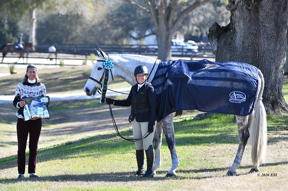 Two women with a gray horse, one holding a ribbon and the other holding the horse's reins, outdoors in a park or equestrian setting.