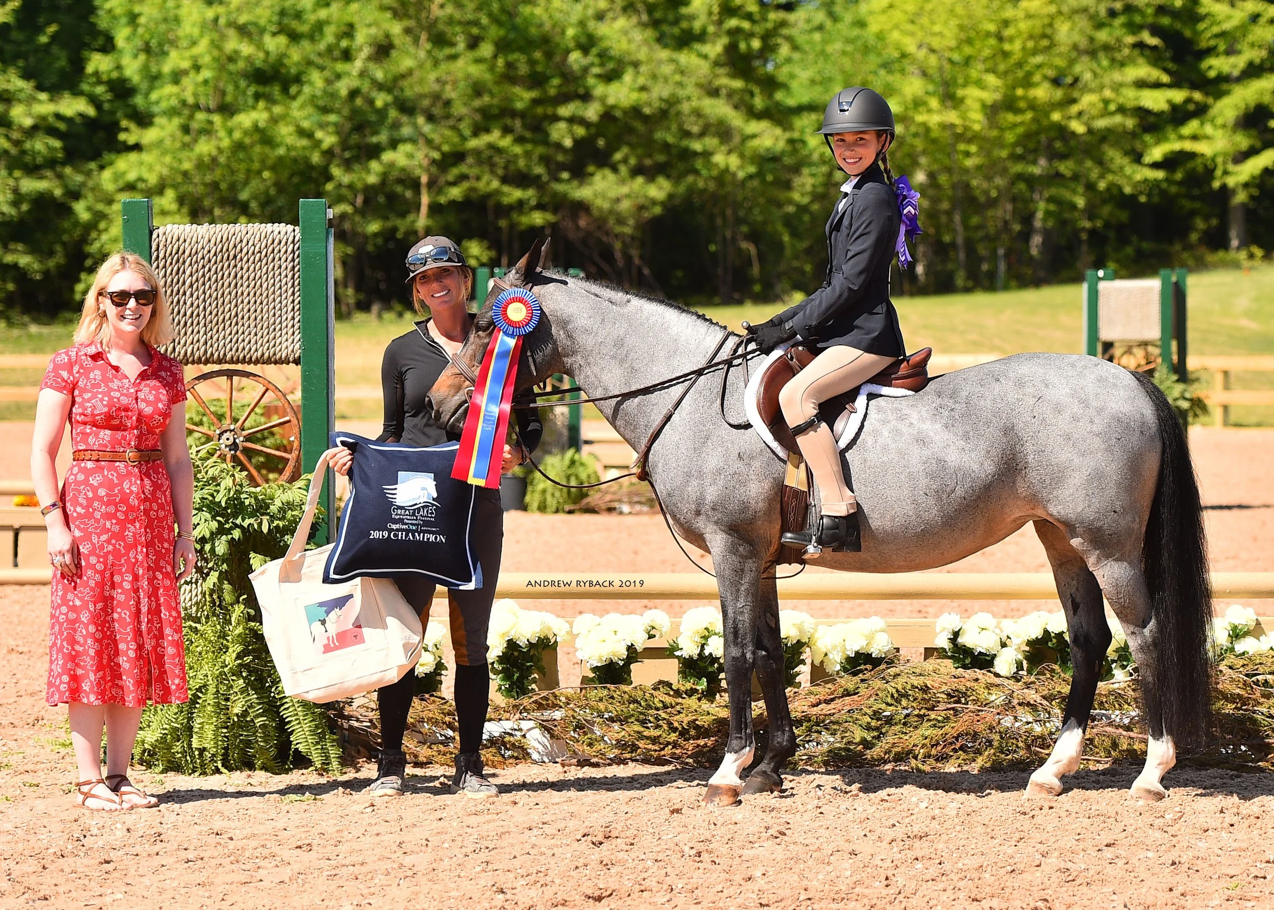 A young girl on horseback at an equestrian competition, receiving a blue ribbon. Two women stand nearby, one in a red dress and sunglasses, the other in black, holding a banner. The scene is outdoors with trees and a sandy riding arena in the backgro