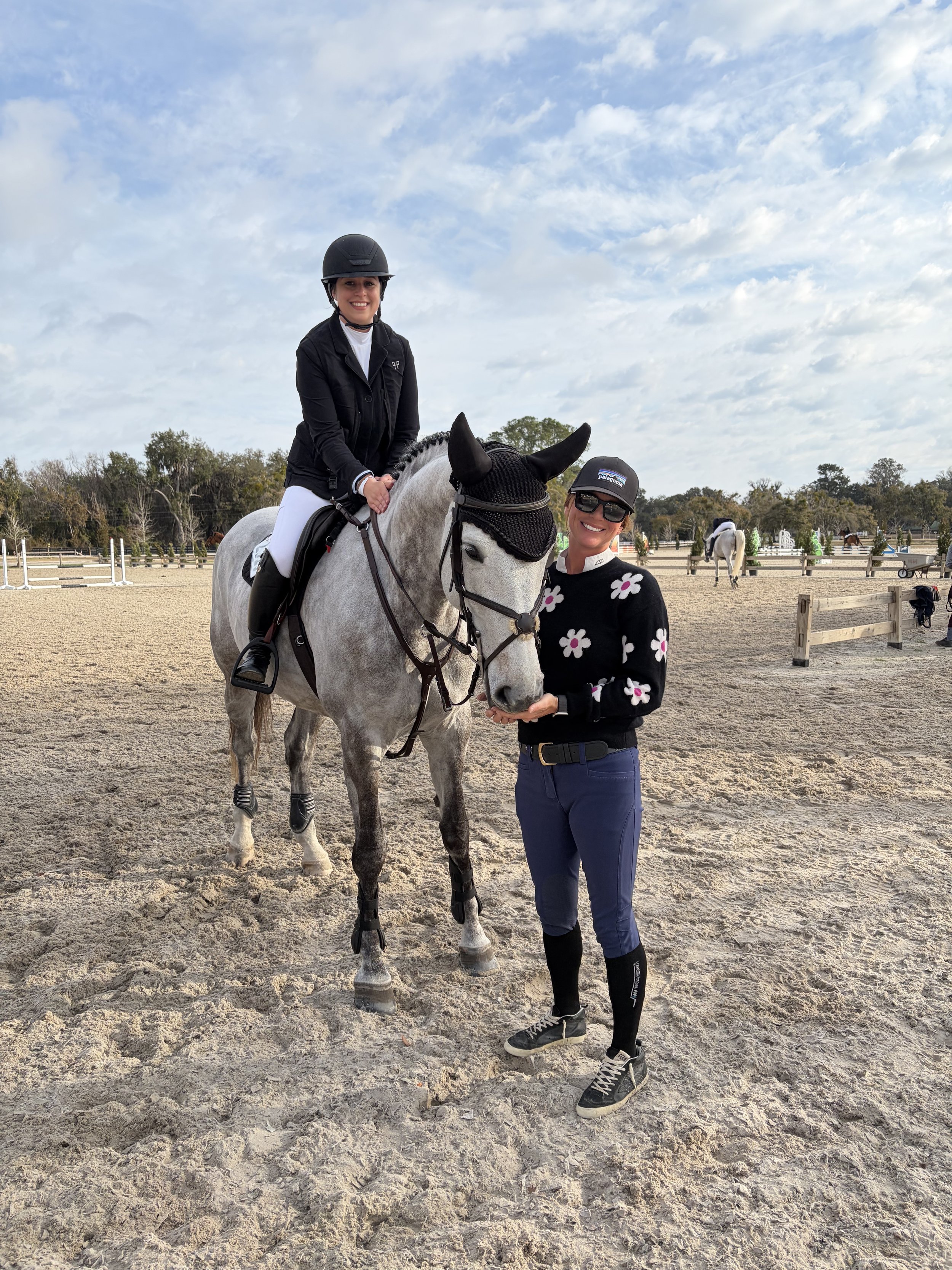 A woman standing next to a white horse with a black mask, while a girl sits on the horse, all smiling in an outdoor riding arena with a blue sky and trees in the background.