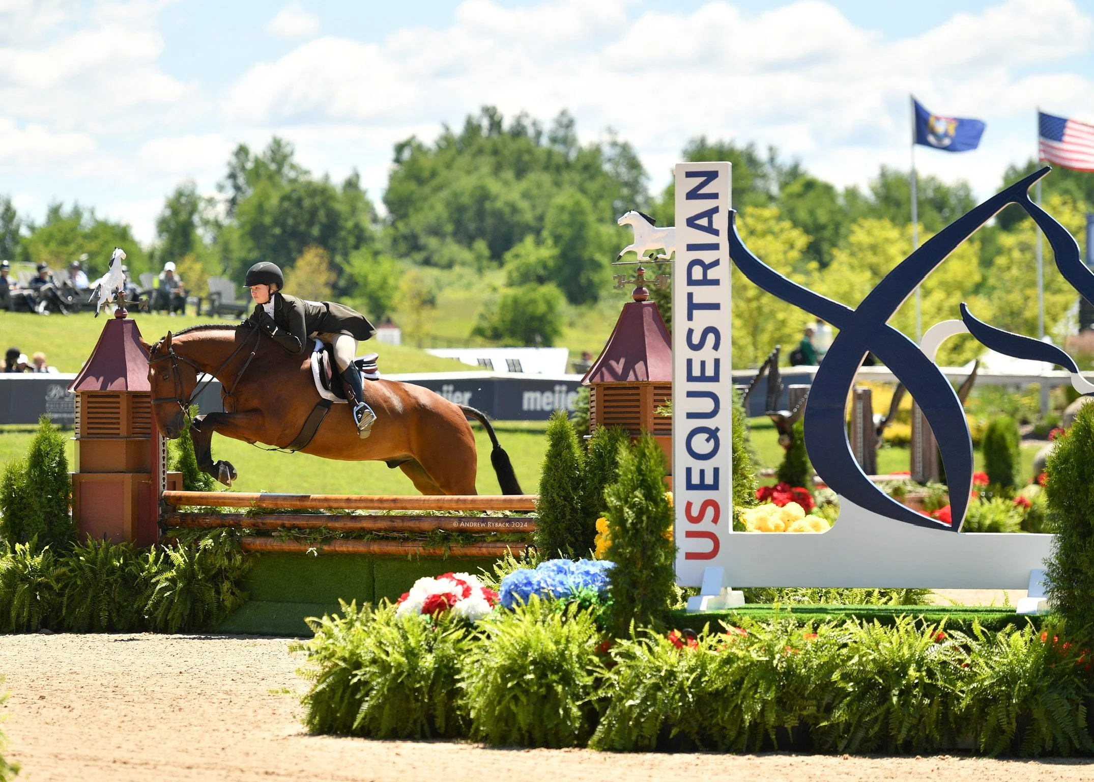 A rider in equestrian attire riding a horse over a jump at a show. The jump includes a blue sign with the word "QUESTRIAN" and decorative elements, with American and regional flags in the background under a partly cloudy sky.