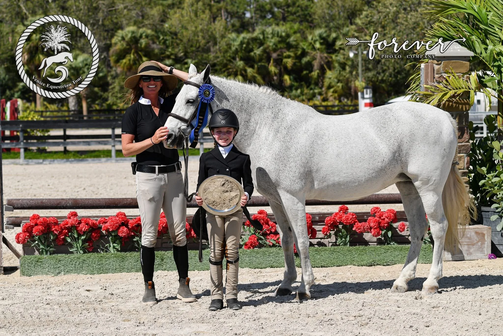 A woman and a girl standing next to a white horse decorated with a blue ribbon at an equestrian event. They are smiling, and the woman is wearing a large sunhat and sunglasses. The girl is dressed in equestrian attire, including a helmet and riding b