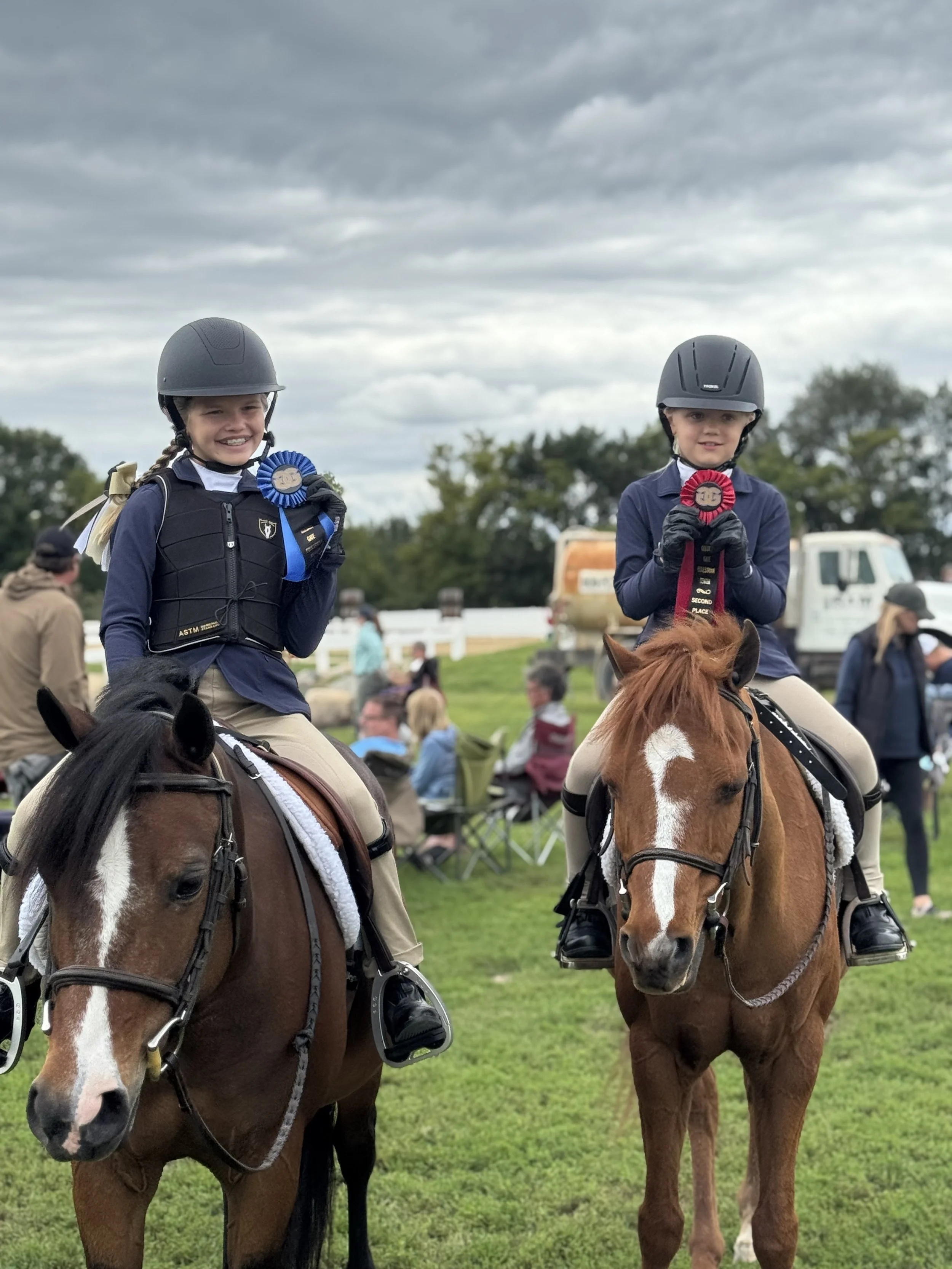 Two young girls in riding gear, including helmets, sitting on horses, holding blue and red ribbons, at an outdoor event on a cloudy day with people and trees in the background.