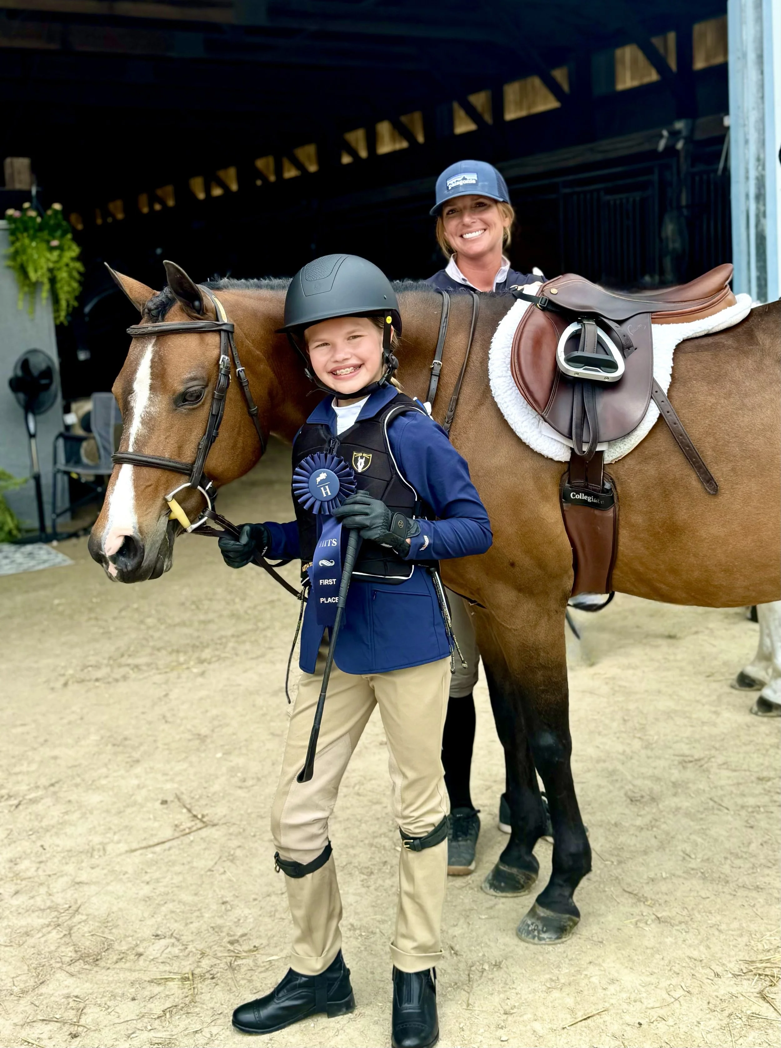 Smiling woman and young girl with riding helmets, standing beside a horse with a saddle and bridle in a stable or riding arena.
