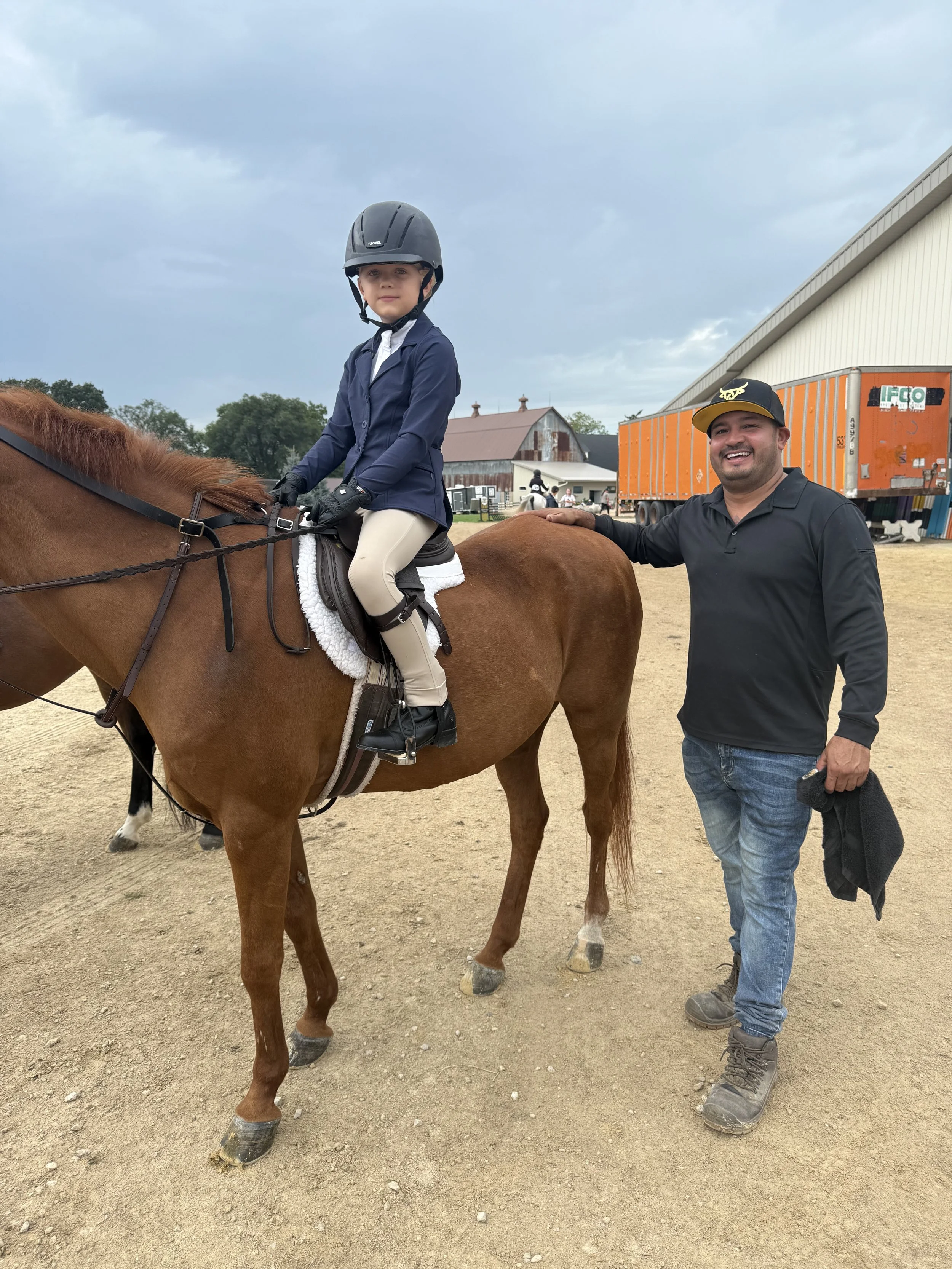 A young girl wearing a helmet and riding a brown horse, with a smiling man standing beside them outdoors on a dirt surface. There is a barn and several other people in the background, with a cloudy sky overhead.