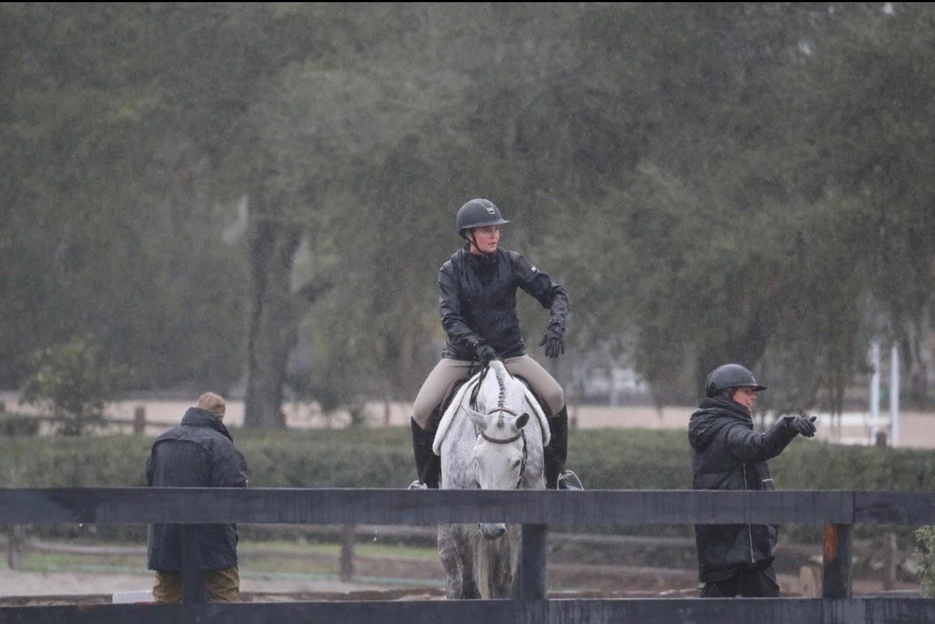 A woman riding a white horse during a rainy day at a horse riding facility, with two men dressed in black jackets and helmets standing nearby.