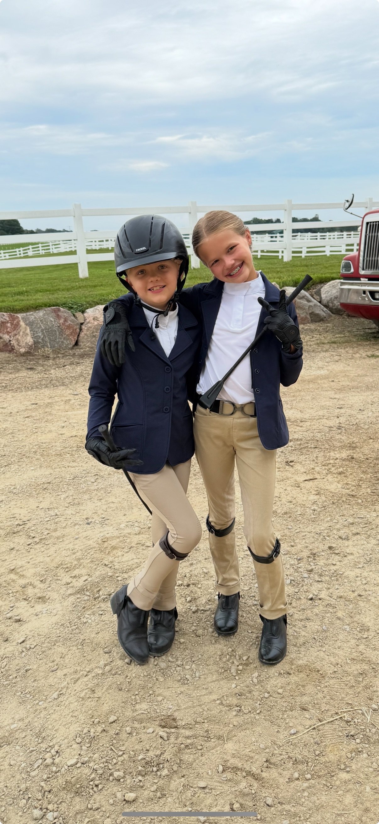 Two young female equestrians in riding attire, standing outdoors on a dirt path, smiling and posing with their arms around each other, with a white fence, green grass, and a red truck in the background.