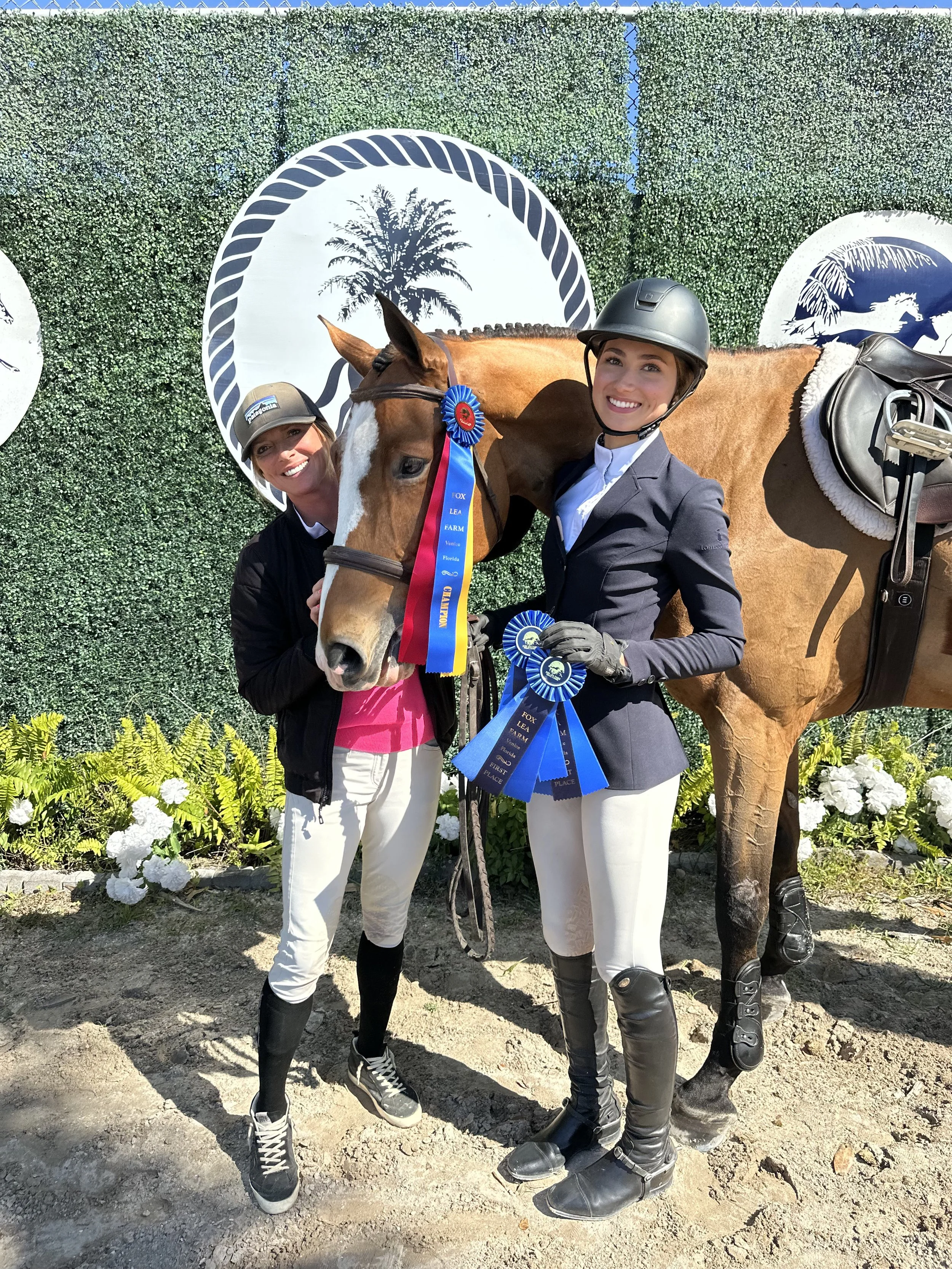 Two women and a horse with ribbons and trophies in an outdoor setting with a decorative green hedge and white flowers in the background.