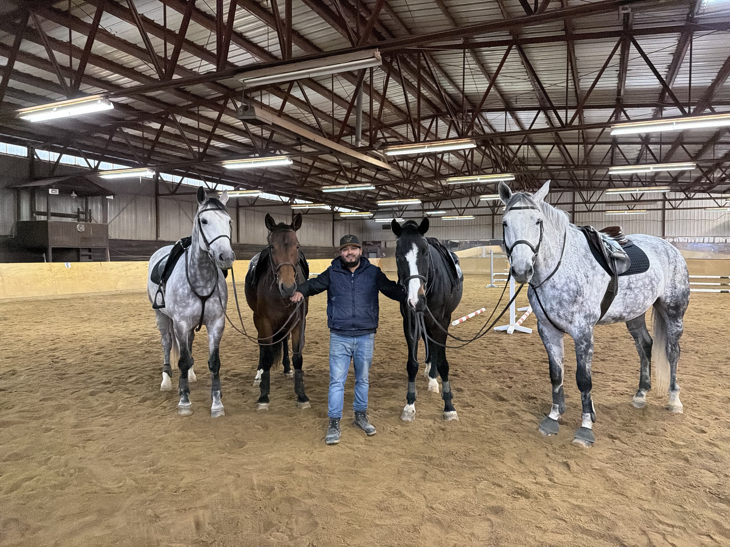 A man is standing in an indoor equestrian arena, holding the reins of four horses. The arena has a dirt floor, a high metal roof, and artificial lighting. The man is smiling and wearing a dark jacket and a cap.