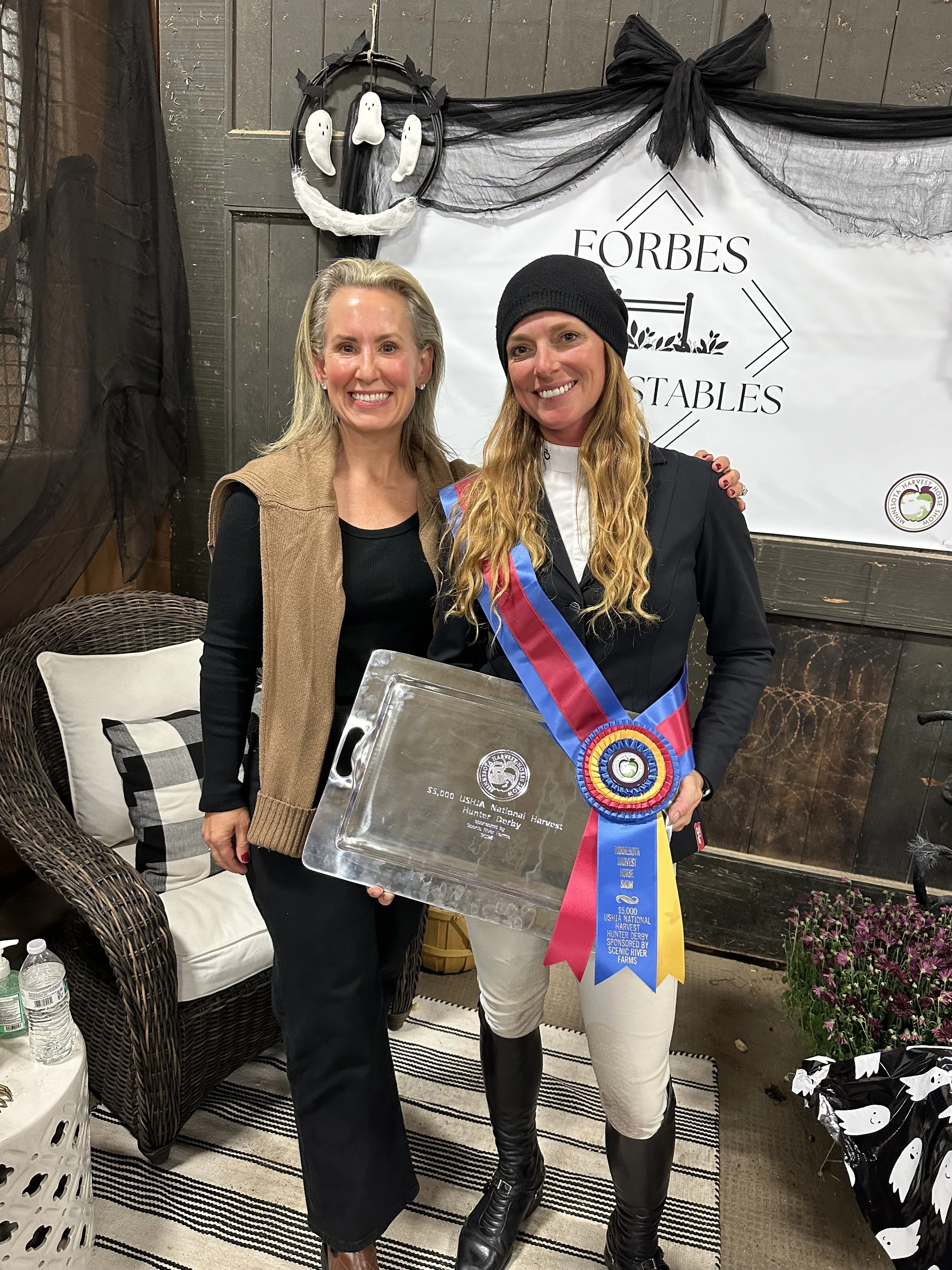 Two women smiling, one holding a large silver tray with an inscription, standing in front of a black and white banner that reads 'Forbes' and 'Stables'. The woman on the right is wearing a black outfit with a sash and a medal ribbon, possibly a ridin