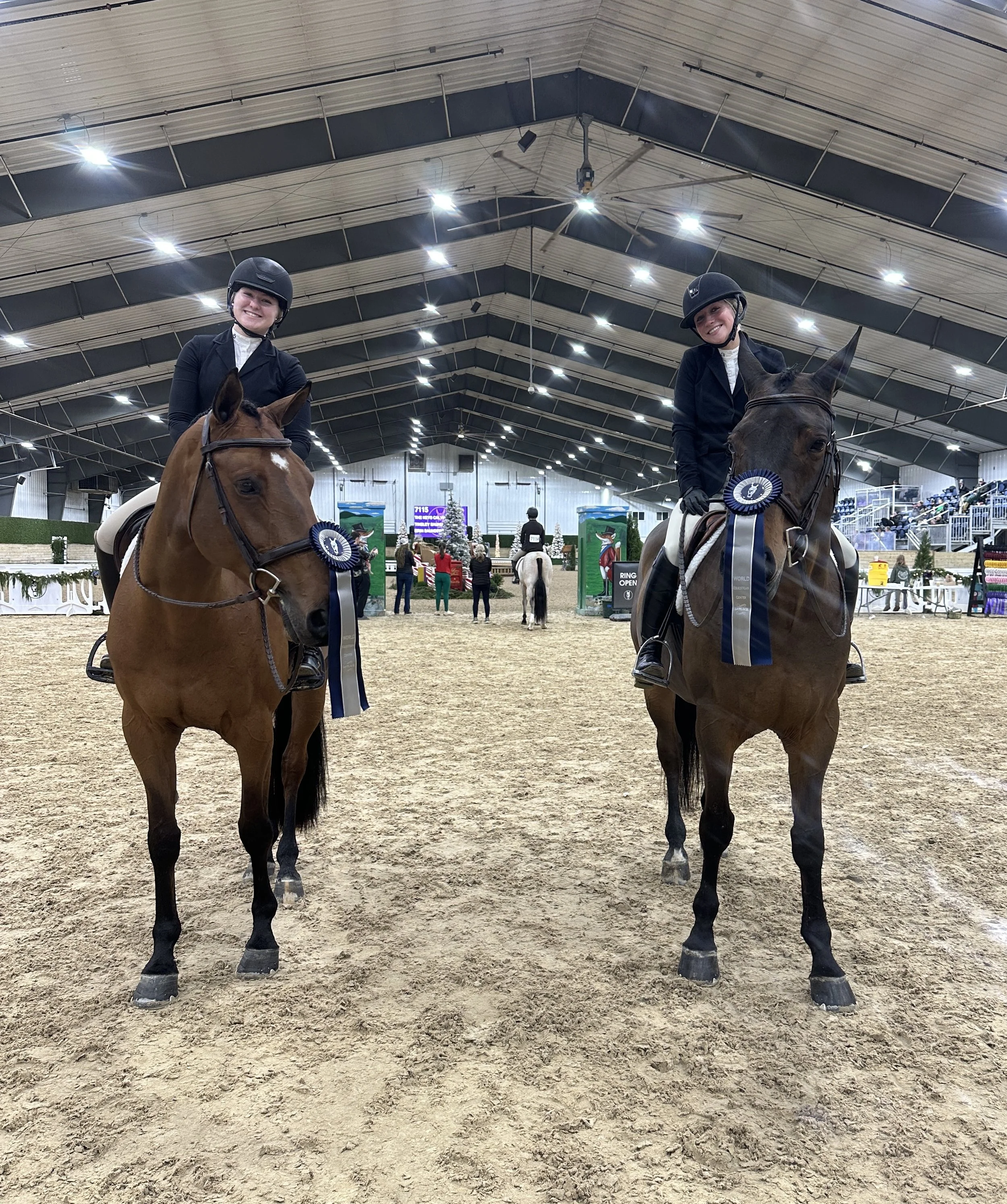 Two smiling women dressed in black riding attire and helmets, sitting on brown horses with ribbons, inside an indoor equestrian arena with a high curved ceiling and spectators in the background.