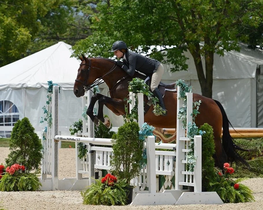 A female equestrian riding a horse and jumping over an obstacle during a show jumping competition in an outdoor arena surrounded by greenery and tents.