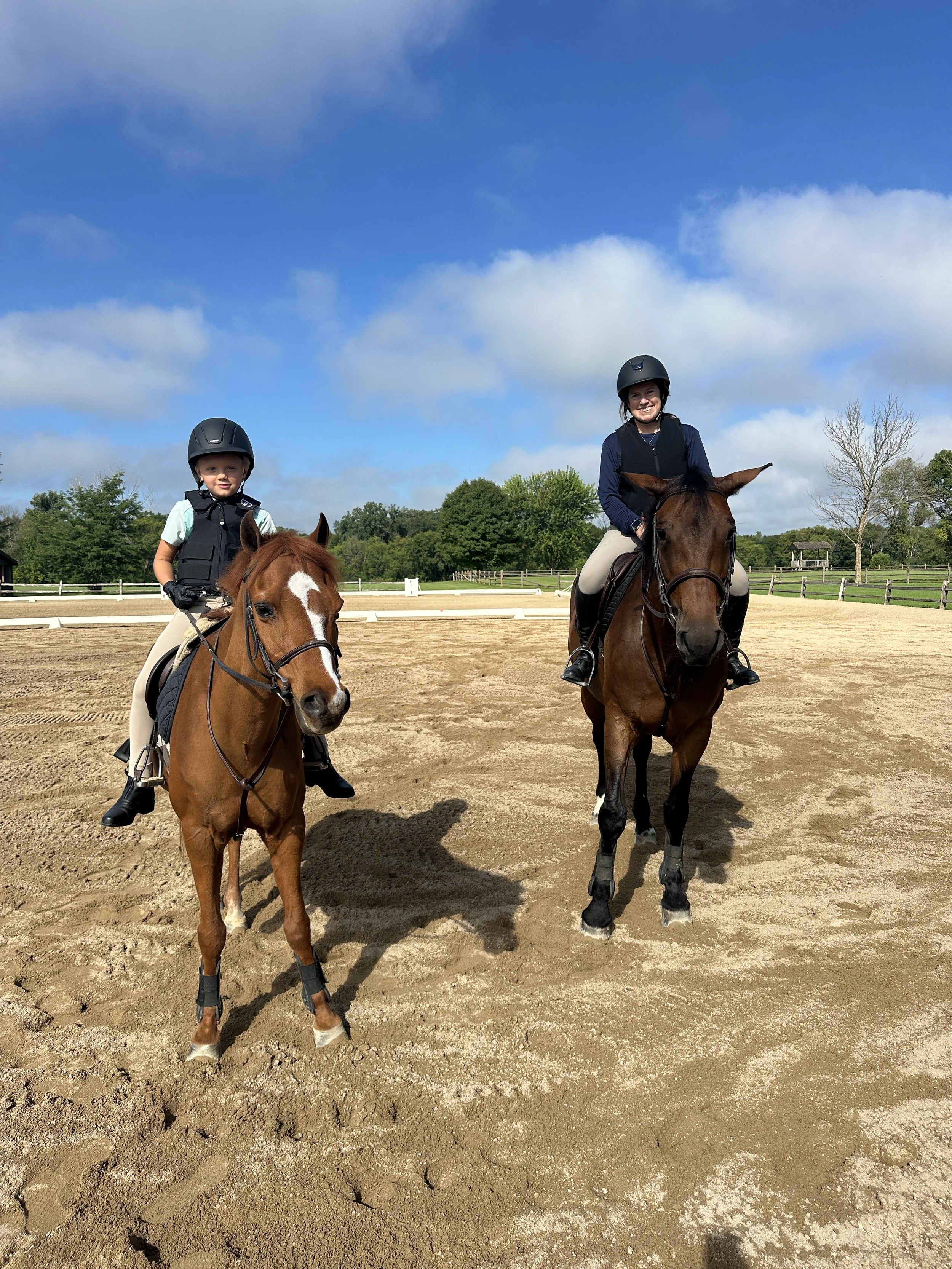 Two children riding horses on a sandy outdoor riding arena, wearing helmets and safety vests, under a partly cloudy blue sky, with green trees in the background.