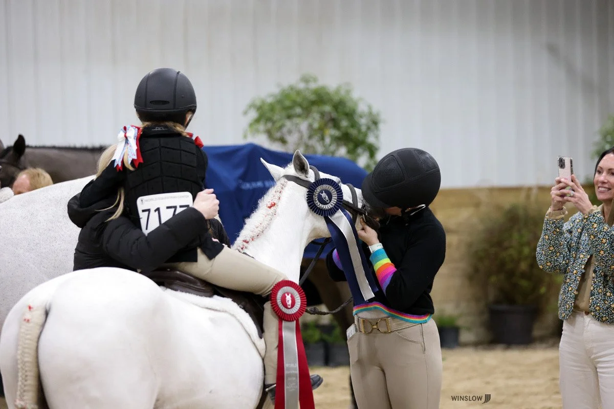 A young girl on a white horse receiving a ribbon from a woman at an equestrian award ceremony, with another woman taking a photo.