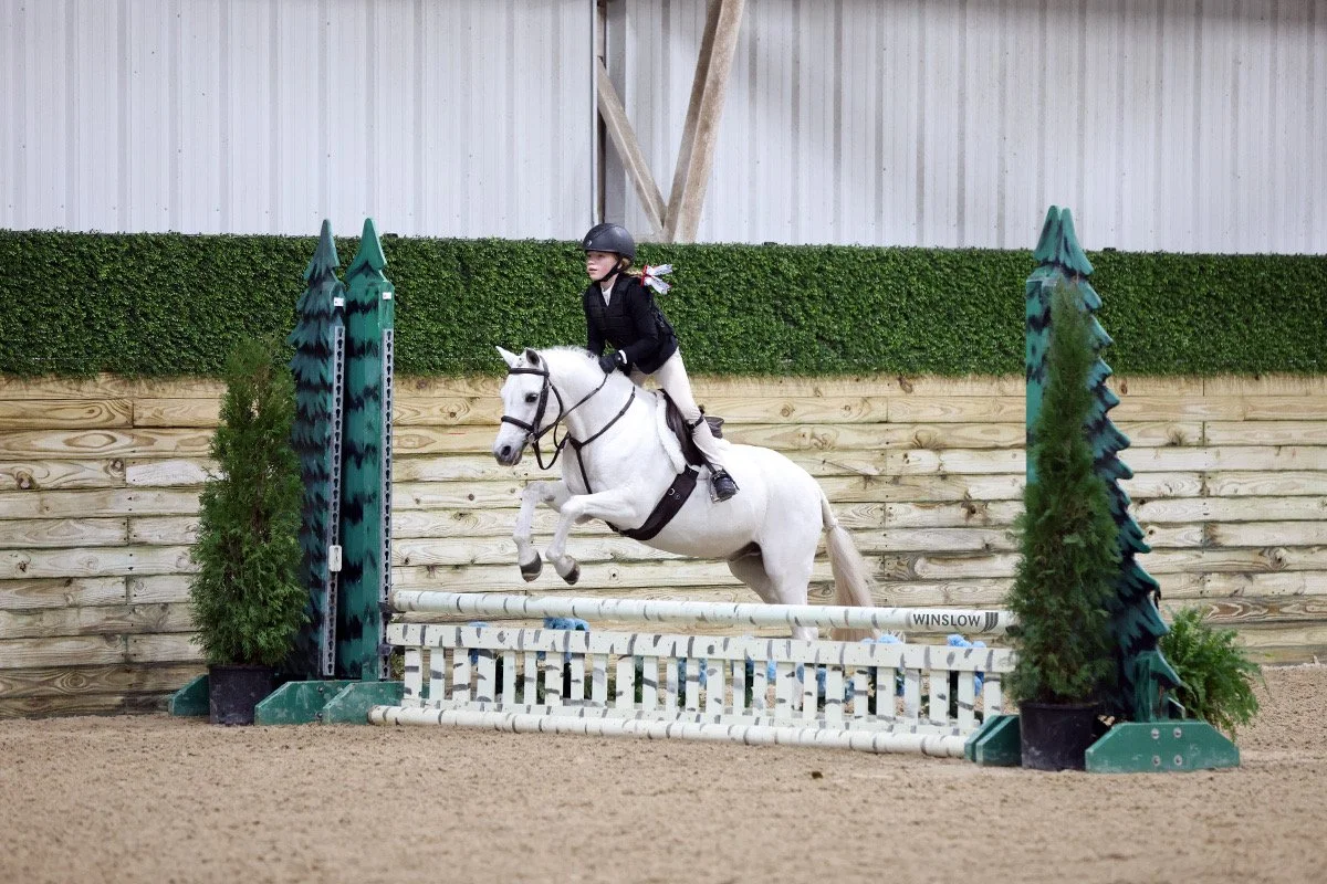 Young girl riding a white horse jumping over an obstacle in an indoor equestrian arena.