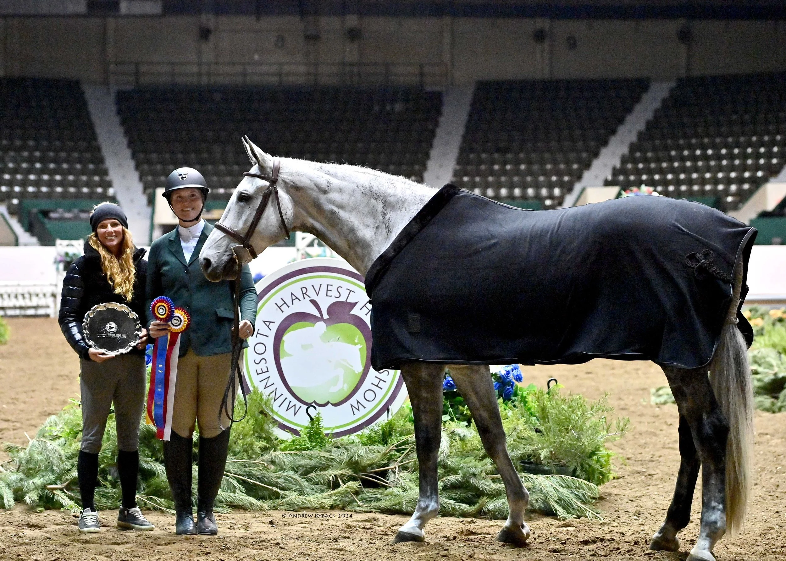 Two women standing with a white horse inside an indoor equestrian arena, holding awards and ribbons, with a round emblem in the background reading 'Somerset Harvest'.