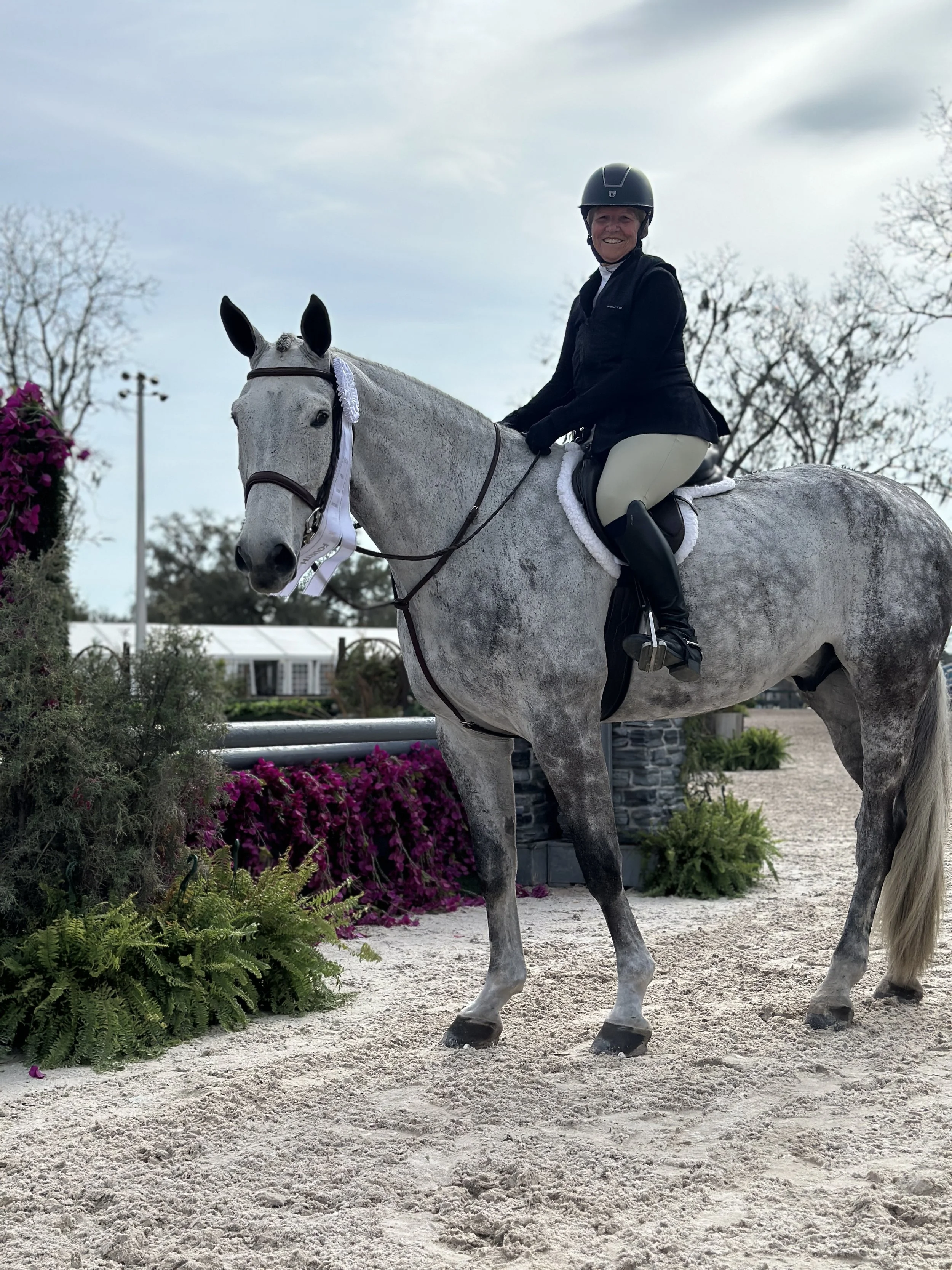 A woman riding a gray horse outdoors, smiling and wearing a helmet, black jacket, beige riding pants, and tall riding boots. The horse has a white ribbon on its bridle, and there are pink flowers and green foliage nearby with cloudy skies overhead.