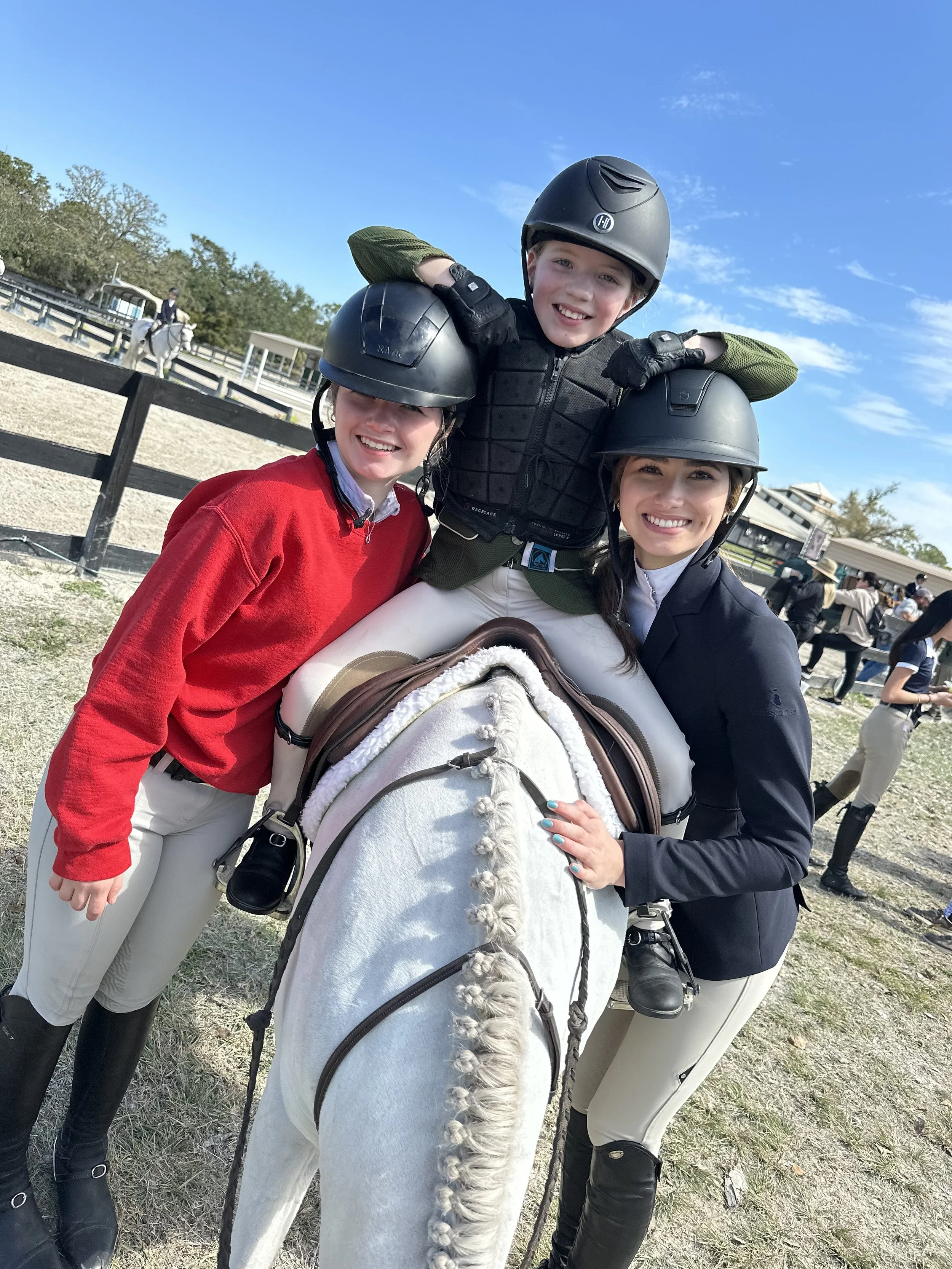 Three smiling children wearing riding helmets posing with a white horse covered with a blanket at an outdoor equestrian center during daytime.