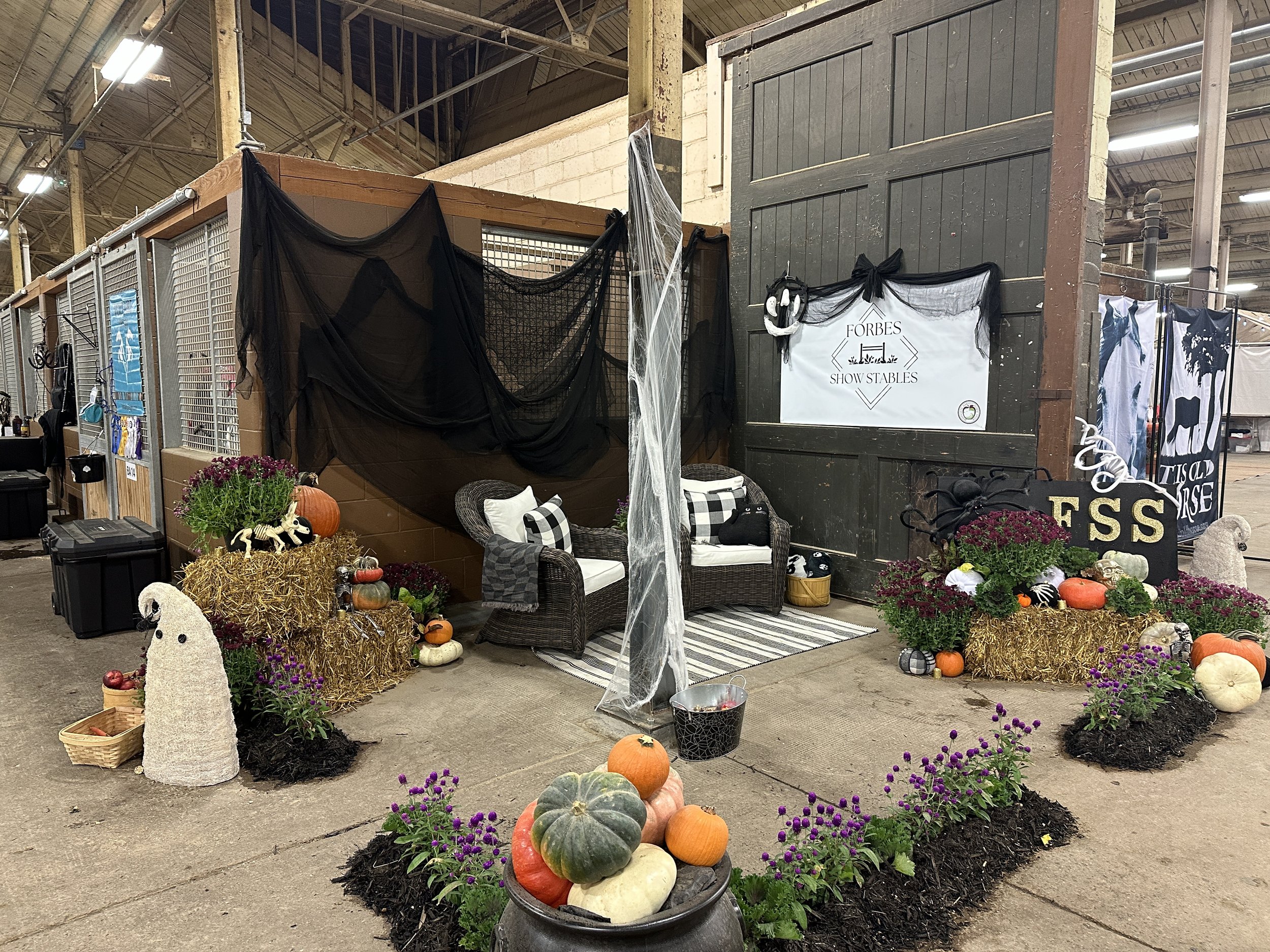 Indoor market stall decorated for Halloween with pumpkins, gourds, flowers, witch and ghost figures, and spider web decorations, with a sign reading 'Forbes Show Stables' on a dark barn-like wall.