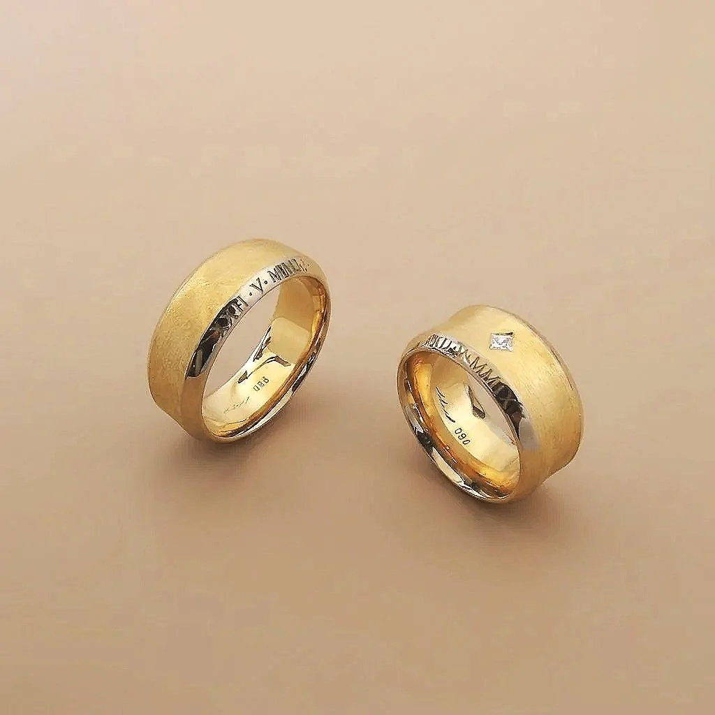 Zwei goldene Eheringe auf hellem Hintergrund, mit eingravierten Namen und einem kleinen Diamanten auf einem der Ringe.