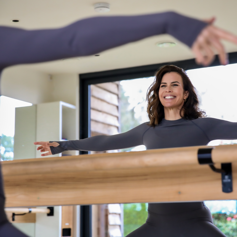 Smiling woman in black workout clothes standing in front of a mirror with her arms extended horizontally during a fitness class, with an instructor partially visible in the foreground.