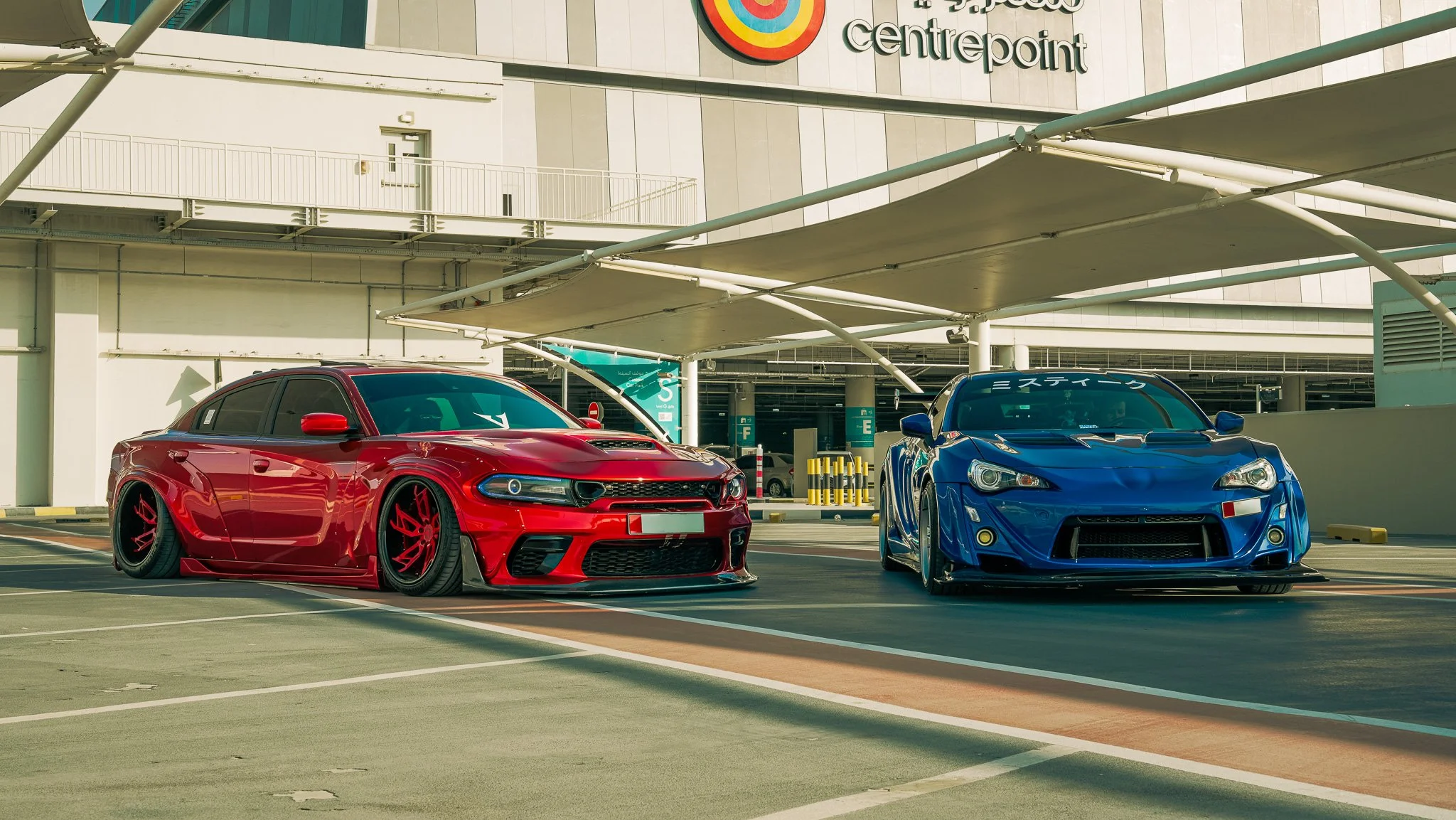 Two sports cars, one red and one blue, parked side by side in an outdoor parking lot at a shopping center called 'Centrepoint'. The background shows a modern building with a large colorful circular sign and a covered area above the cars.