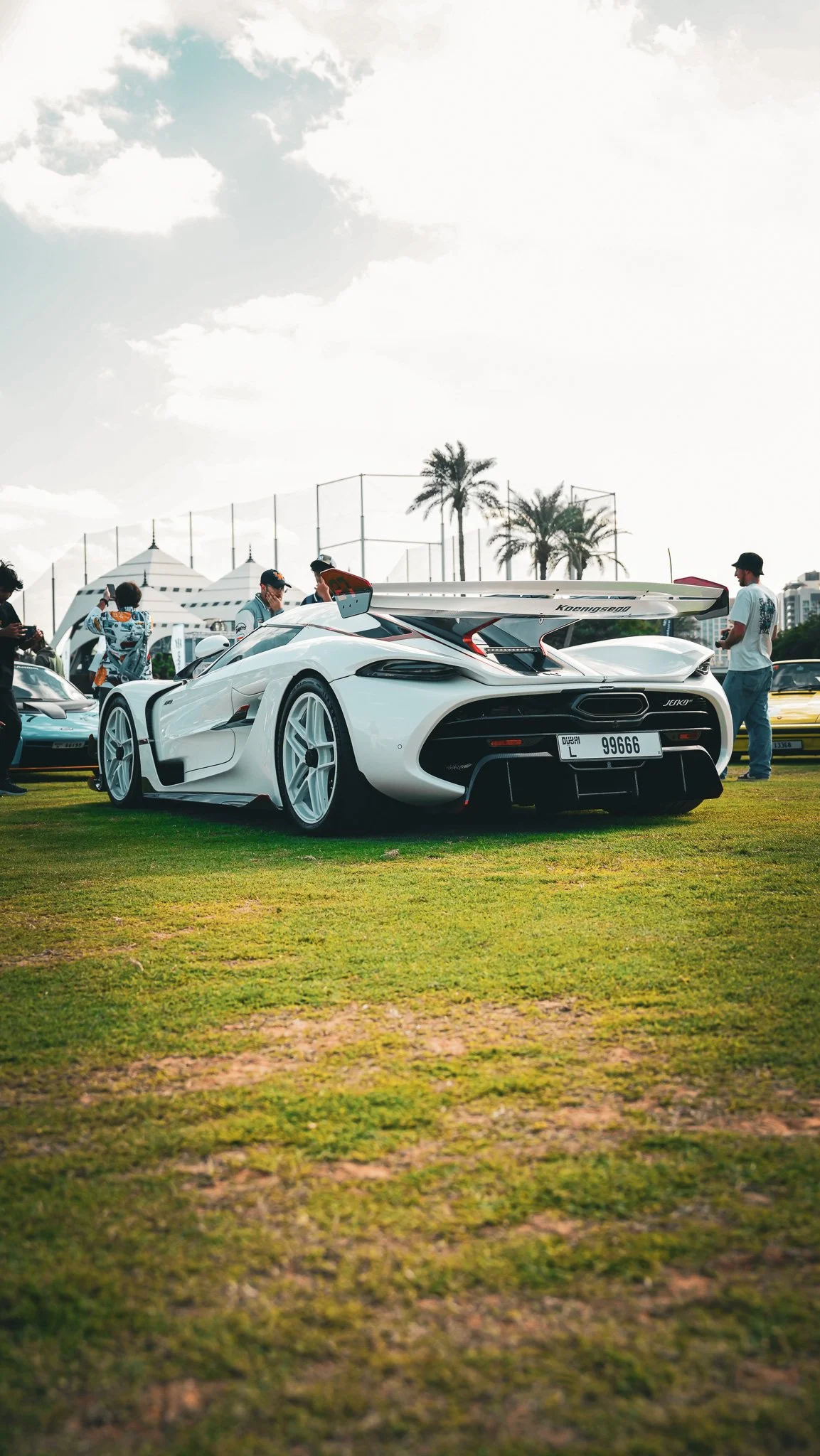 White sports car with a large rear wing on display at an outdoor car event, surrounded by people and palm trees, under a partly cloudy sky.
