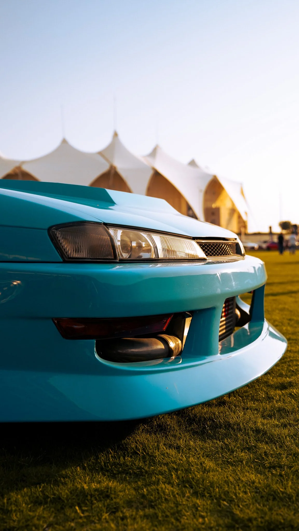Close-up of a vintage light blue sports car with a prominent front bumper and headlights, parked on grass during sunset with tent structures in the background.