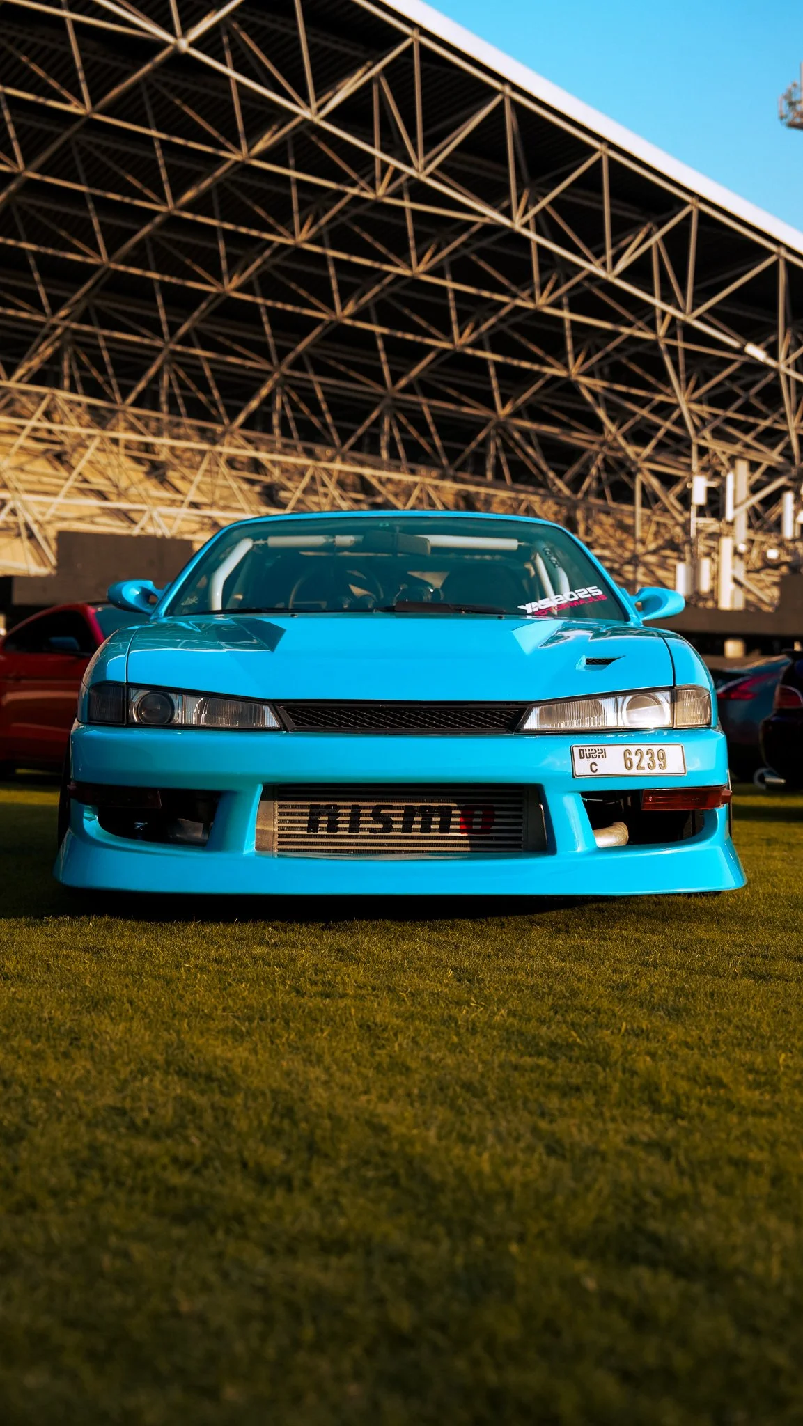 Bright blue Mitsubishi sports car with a custom front bumper and aftermarket grille, parked on grass under a large steel structure.