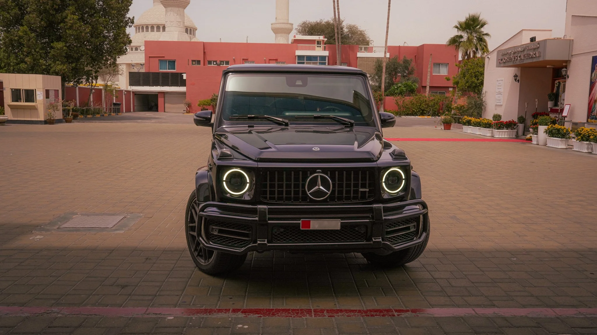 Front view of a black Mercedes-Benz G-Class SUV parked on a paved area in front of pink and beige buildings with a white domed structure and minarets in the background.