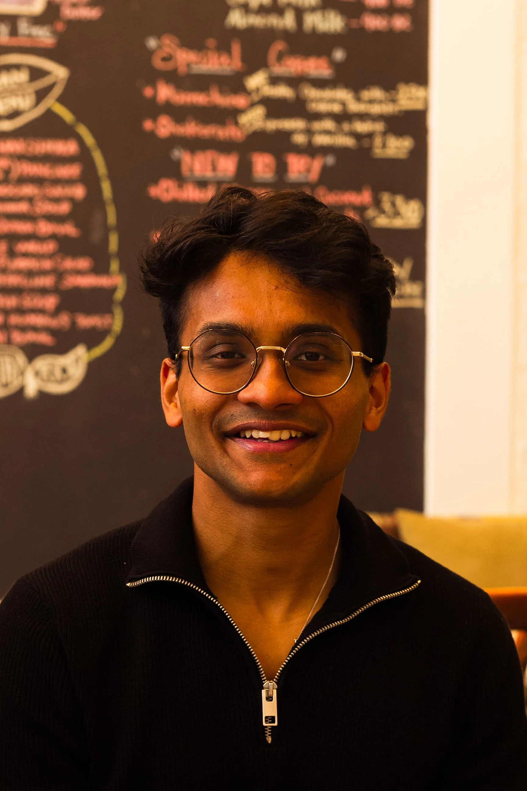 A young man with dark hair and glasses smiling in a restaurant or café with a chalkboard menu in the background.