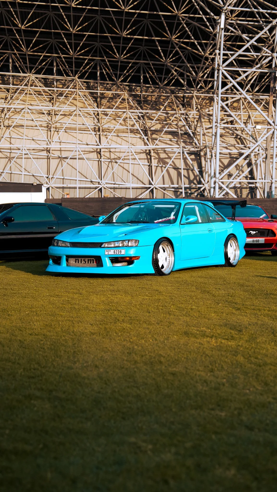 A light blue sports car with a spoiler in front of a lattice-like metal structure, flanked by two other cars on a grassy area.
