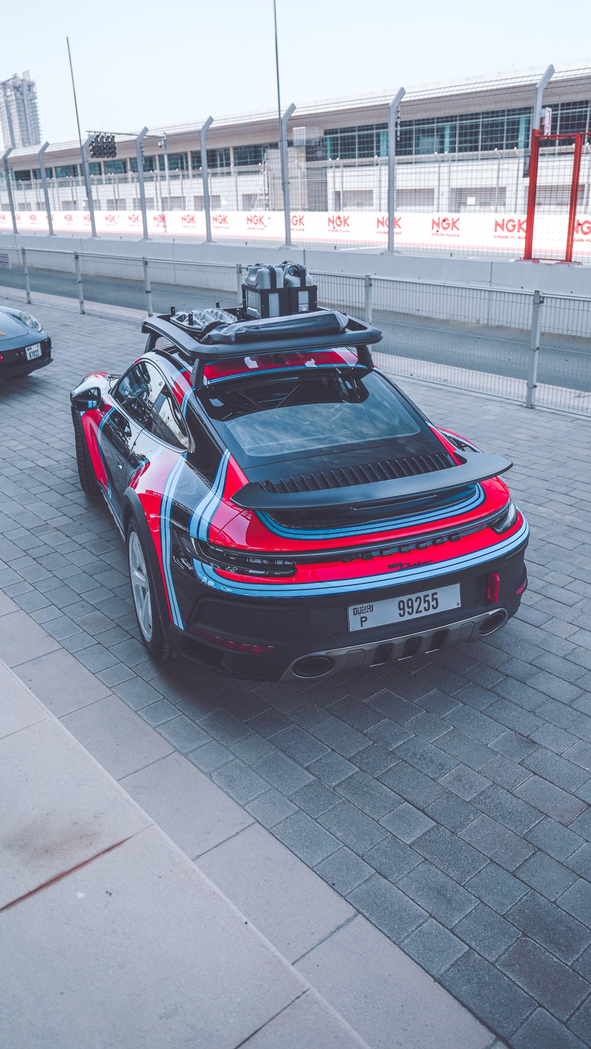 A Porsche sports car with a black, red, and blue racing stripe pattern parked on a brick sidewalk near a race track, with fencing and buildings in the background.