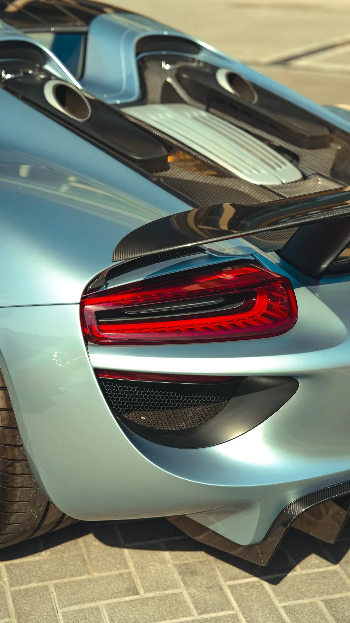 Close-up of the rear section of a silver luxury sports car, showing a red tail light, black carbon fiber diffuser, and part of the rear wheel and tire.
