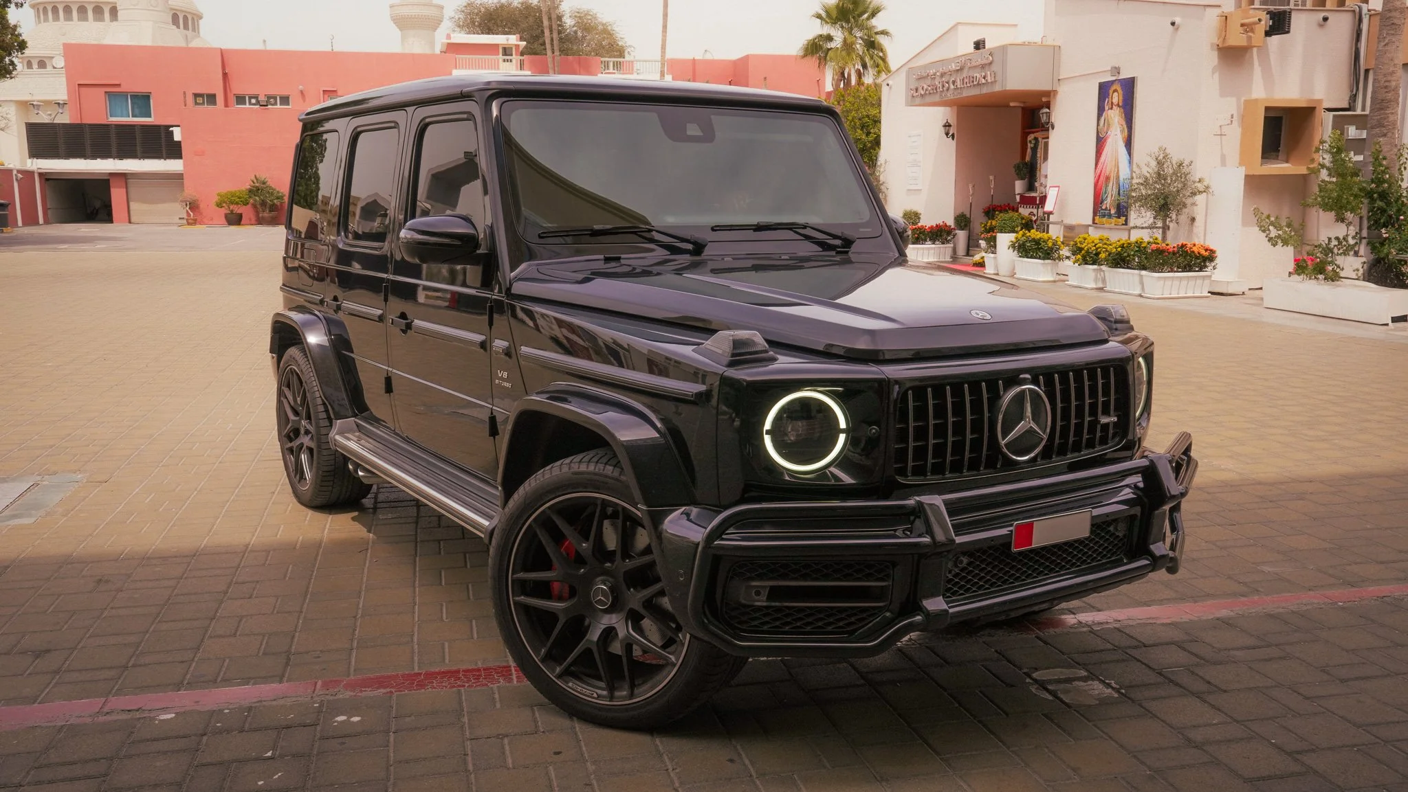 A black Mercedes-Benz G-Class SUV parked on a brick-paved street in front of buildings with flower pots and a religious mural.