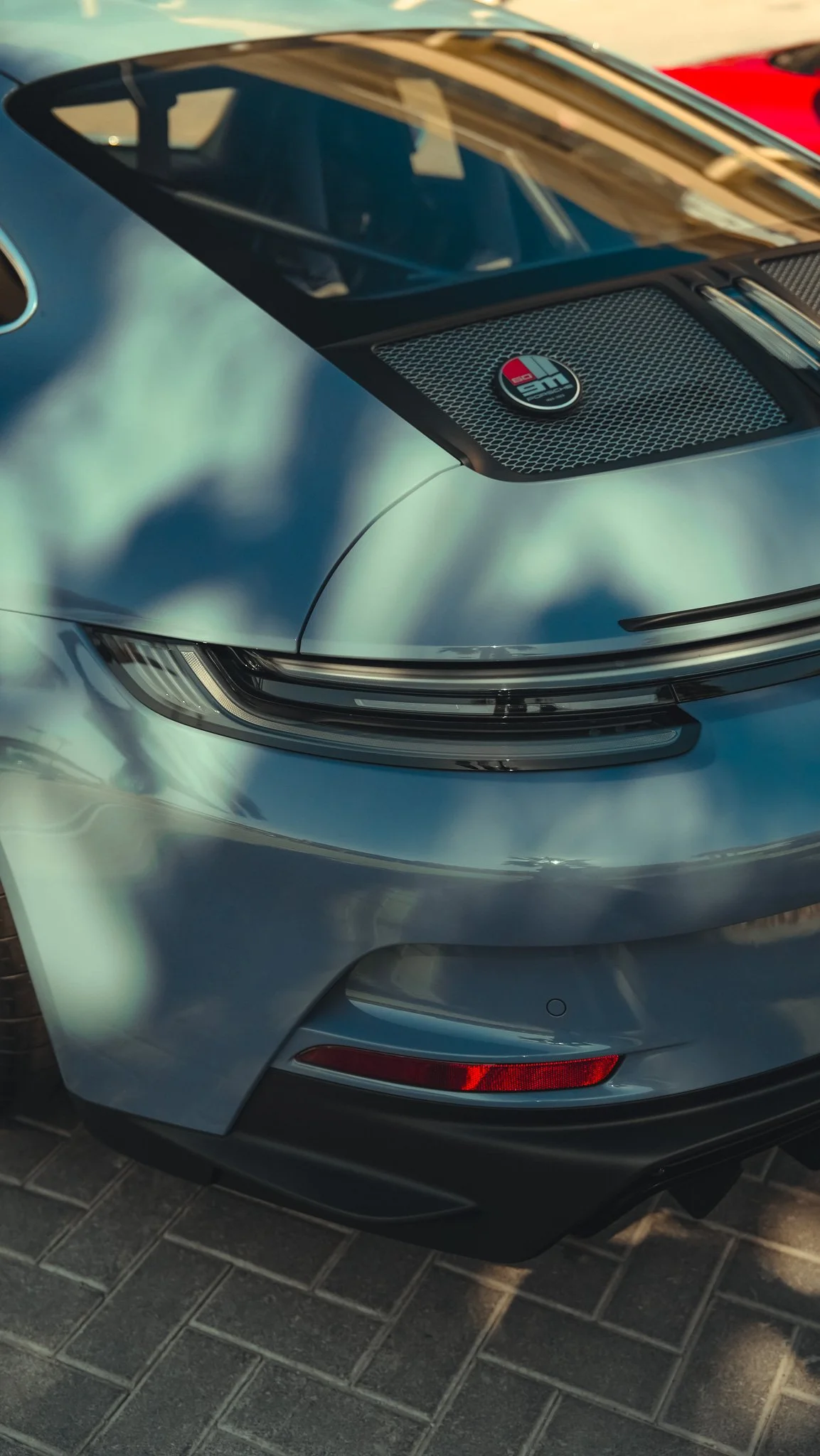 Close-up of the front part of a silver sports car, showing the headlight, hood, and front bumper, parked on a brick pavement.
