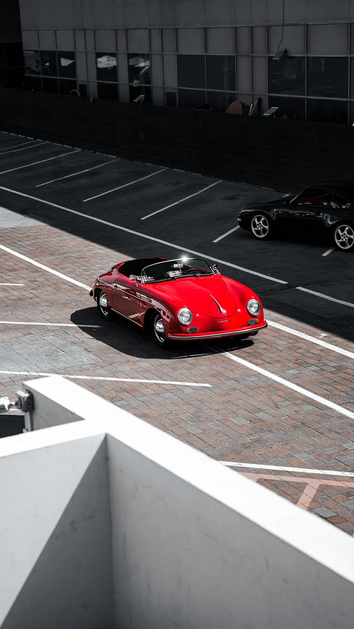 A red vintage Porsche sports car parked in a mostly empty parking lot with dark asphalt and a brick-paved section, adjacent to a modern building with reflective windows.