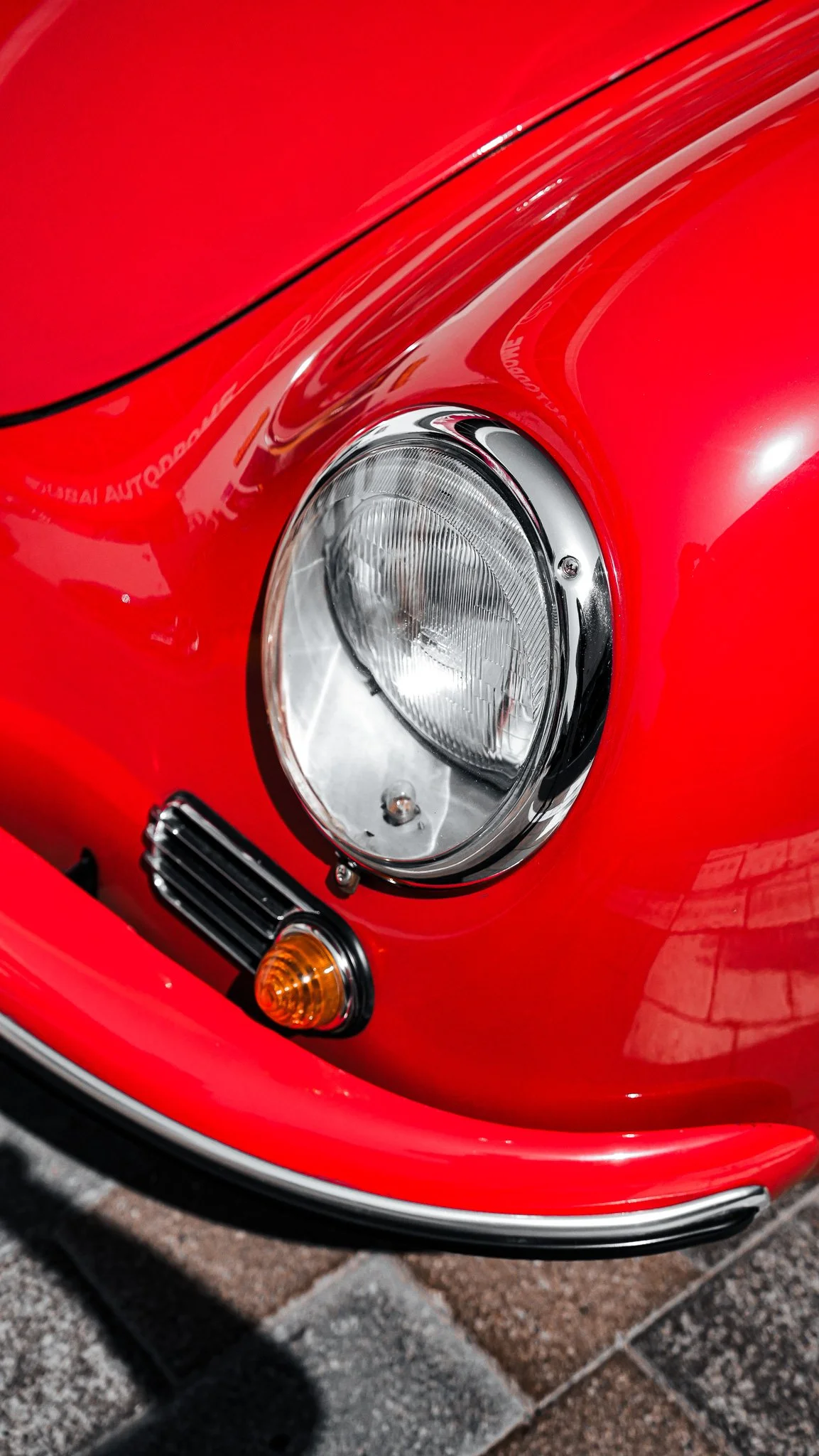Close-up of the front headlight and turn signal of a red vintage sports car on a paved surface.