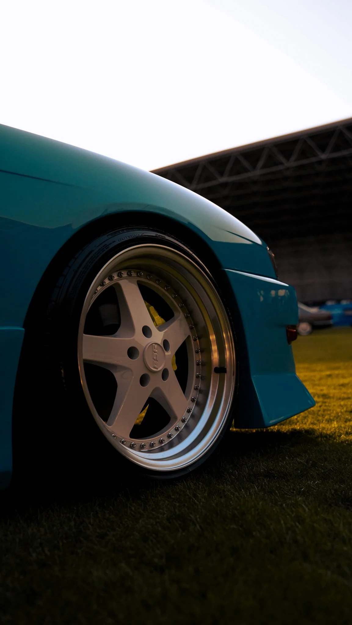 Close-up of a teal car's front wheel and fender at a racing event, with a building structure in the background.
