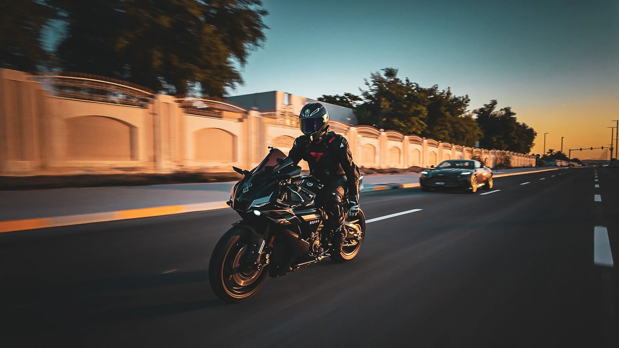 Motorcycle rider in full gear riding on the road during sunset, with cars in the background and trees along the roadside.