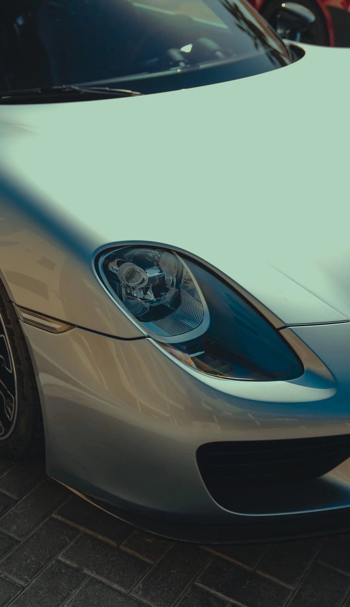 Close-up of the front of a silver luxury sports car parked on a pavement, showing the headlight, part of the bumper, and hood.