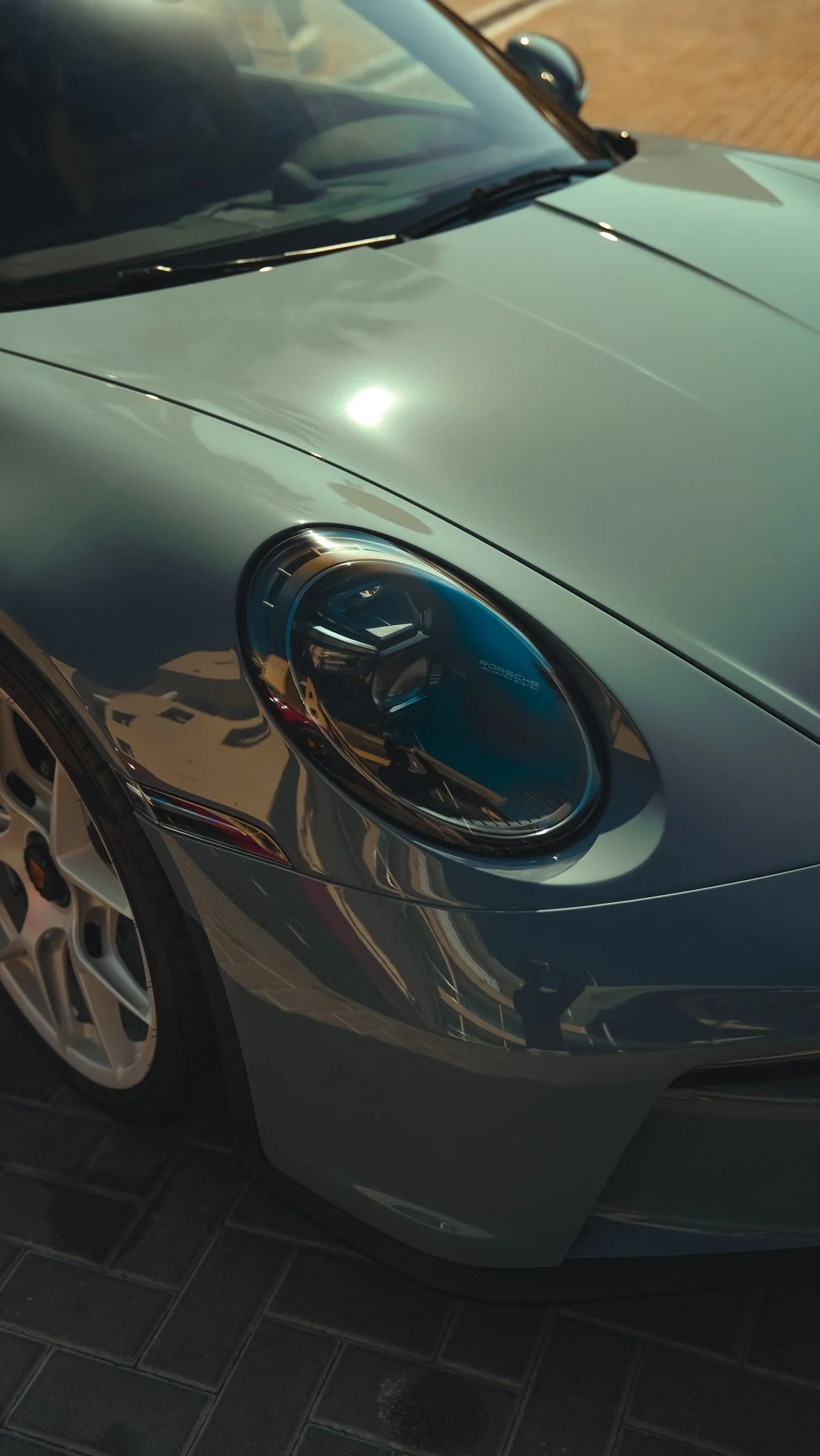 Close-up of a silver Porsche sports car's front left headlight and hood, parked on a brick-paved surface.