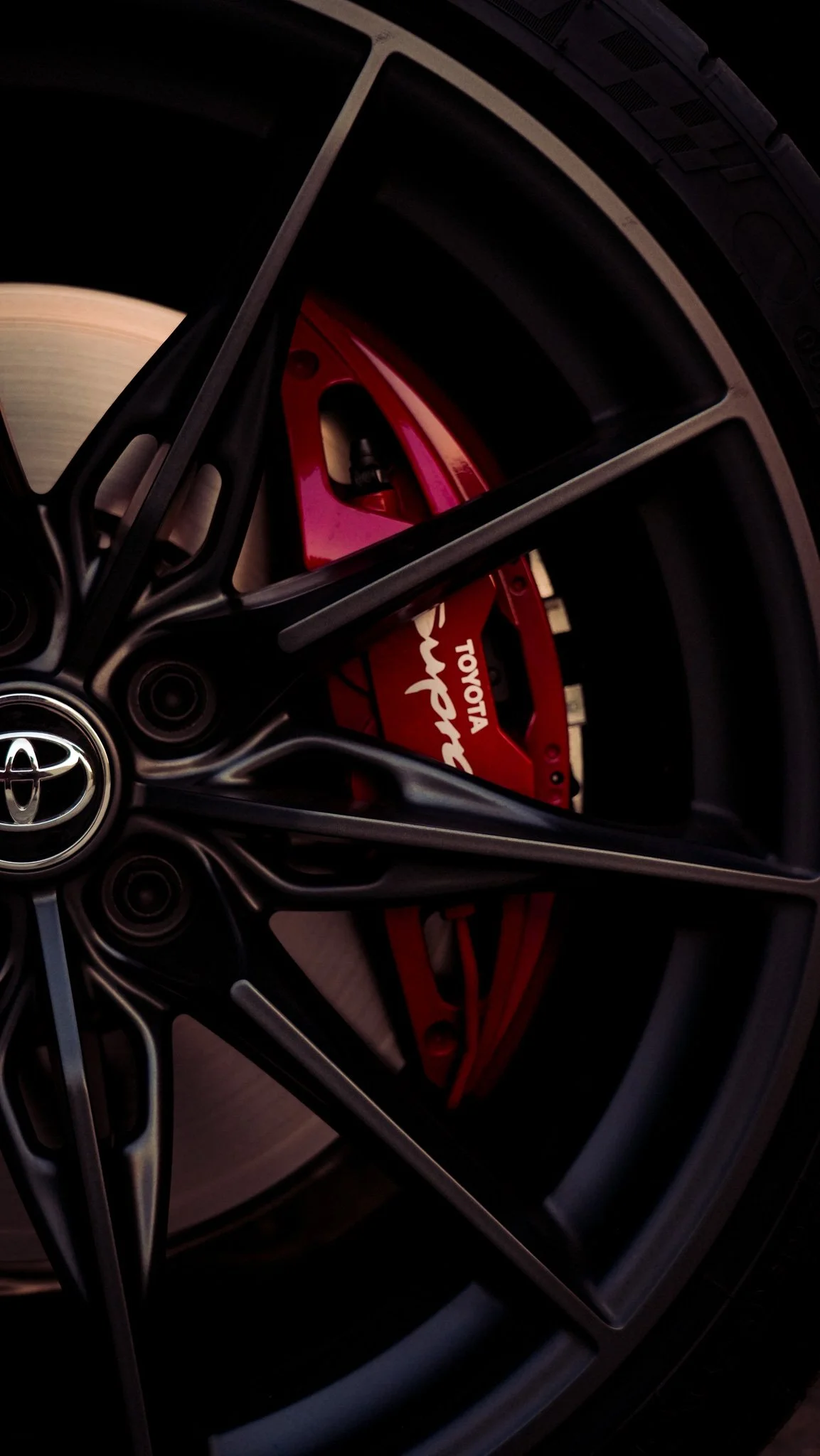 Close-up of a black alloy wheel with a red brake caliper that has the Toyota logo and engraved words. The wheel design features multiple spokes and is partially visible.