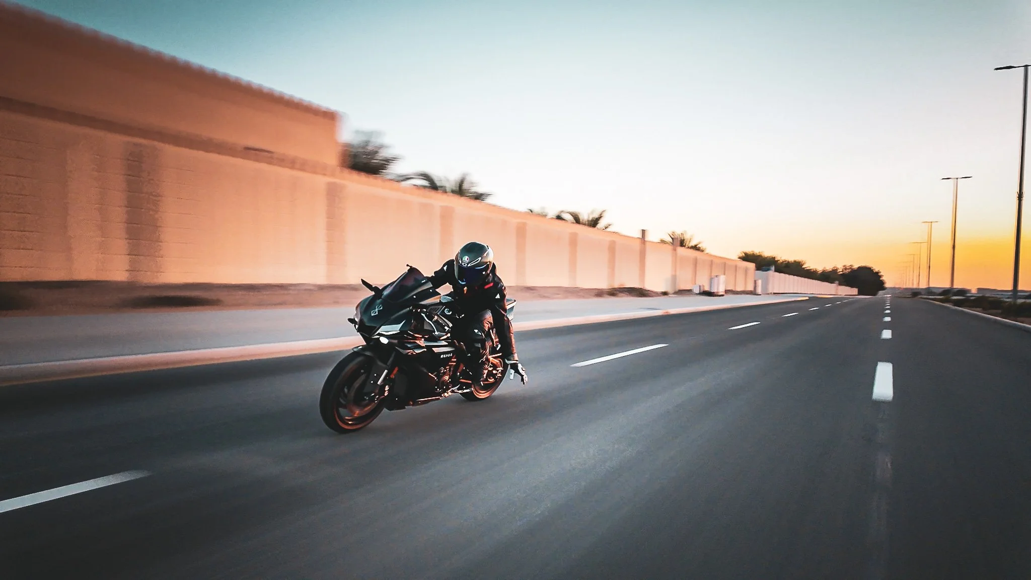 A motorcyclist riding a black sports bike on an empty highway at sunset, with a tall beige wall on the left and streetlights on the right.