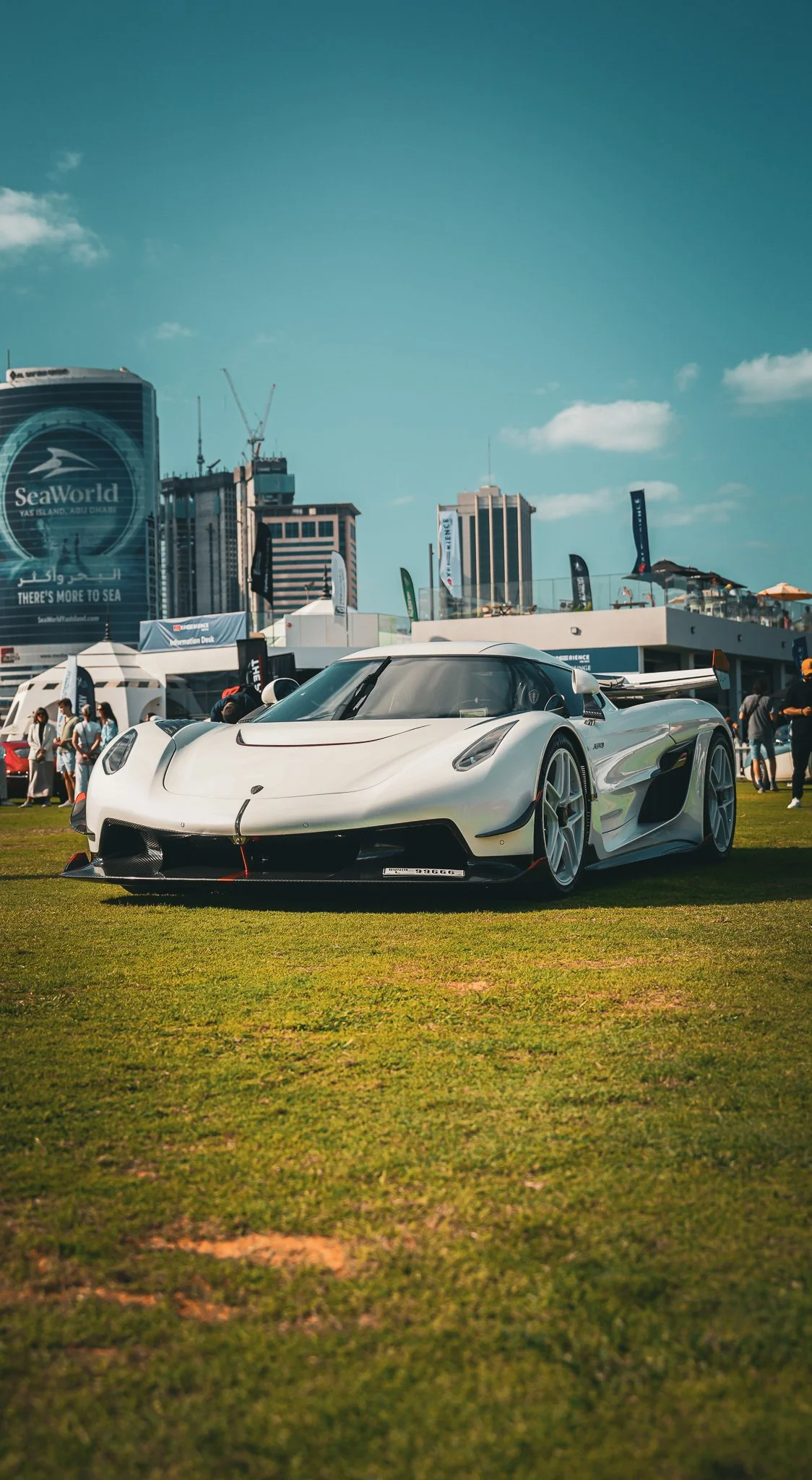 A white sports car on a grassy field at a car show with people and tents in the background, under a clear blue sky.